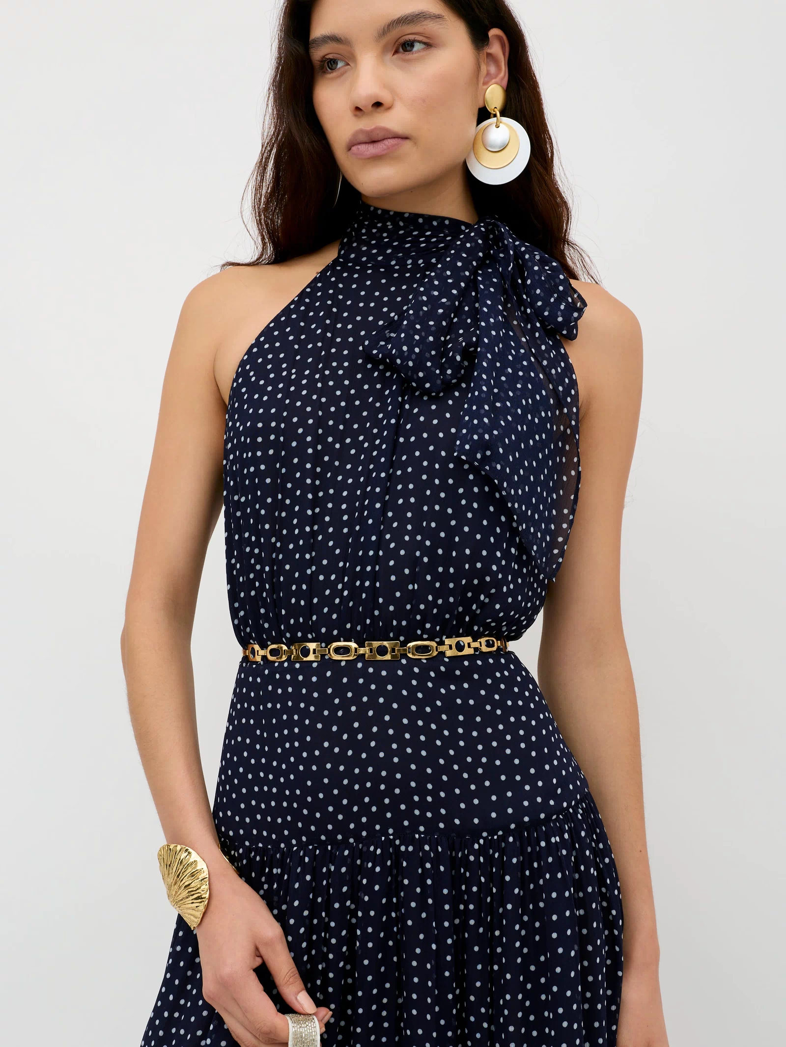 A woman wears the Abi - Mini Navy Polka Dot dress, a sleeveless georgette design with white spots, high bow neckline, gold chain belt, statement earrings, and cuff—perfect for special occasions—posed against a plain light backdrop.