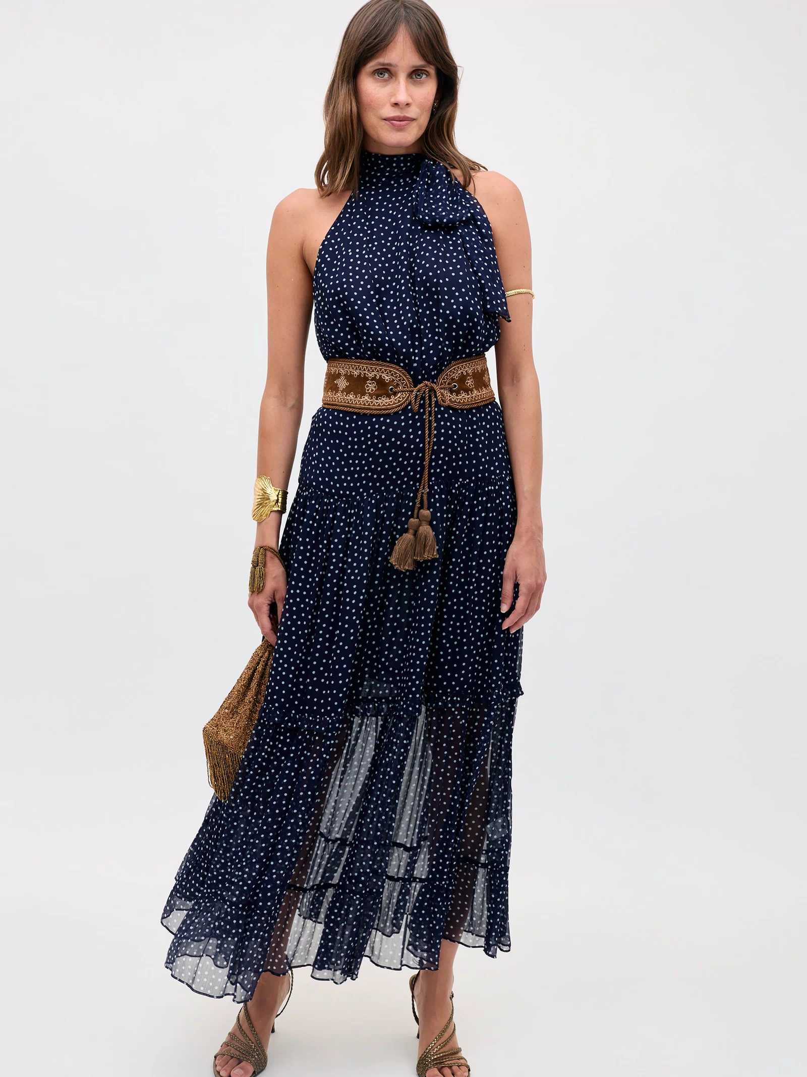 A woman in the Abi - Mini Navy Polka Dot dress, styled sleeveless with a high neckline, accessorized with a brown belt, gold jewelry, tan sandals, and a woven bag, poses against a plain light background.