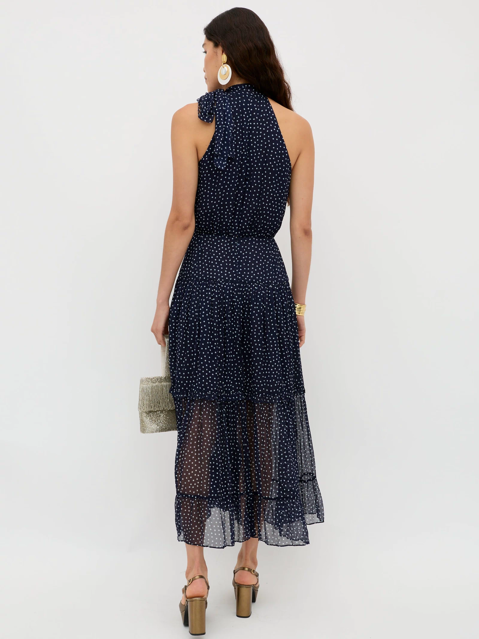 A woman with long dark hair stands facing away, wearing the Abi - Mini Navy Polka Dot dress with heeled sandals. She holds a light woven handbag and accessorizes with large hoop earrings and a gold watch against a white background.