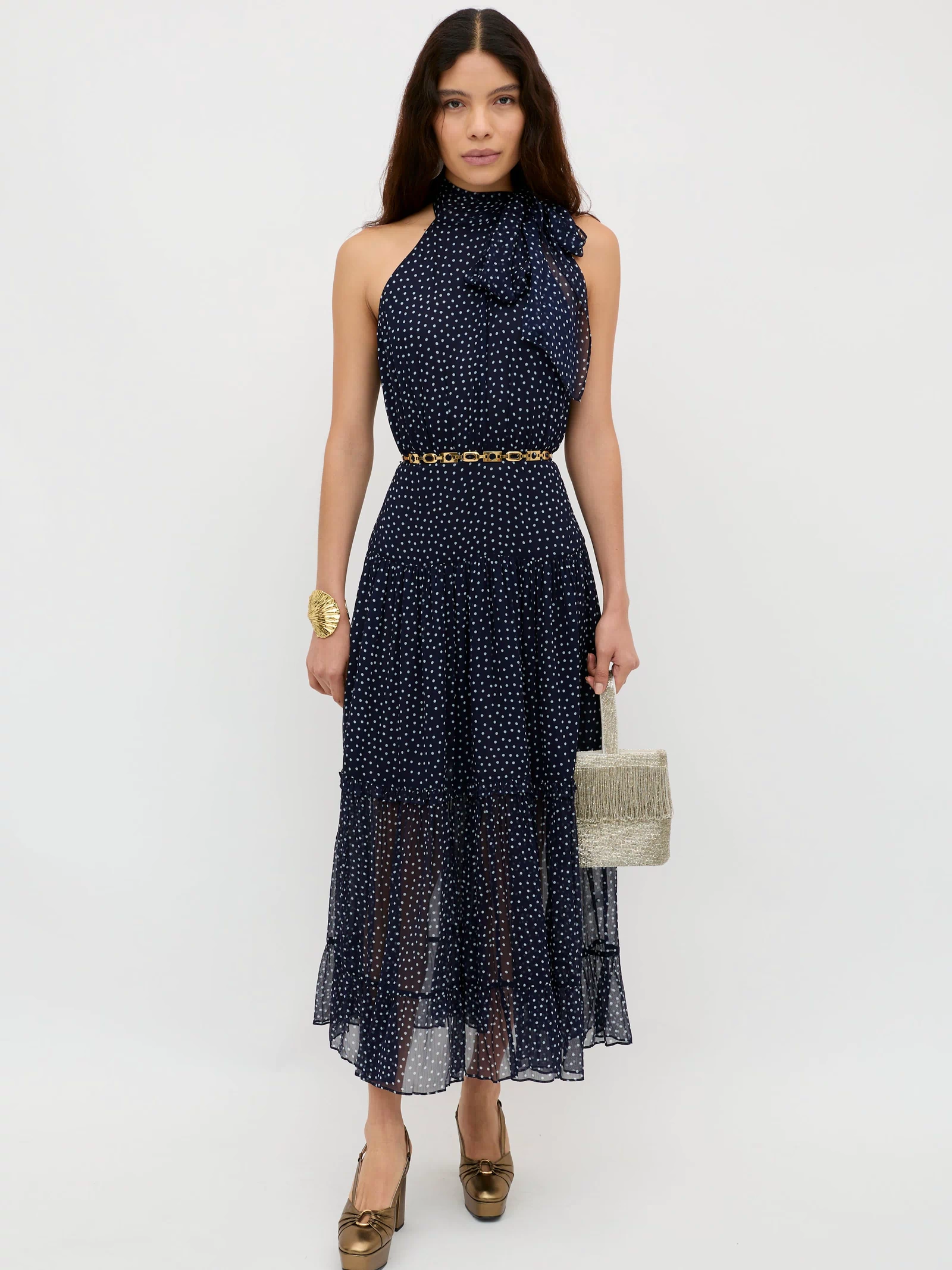 A woman poses against a white background wearing the Abi - Mini Navy Polka Dot georgette dress with halter neckline and ruffle detail, styled with a gold belt, gold heels, gold bracelet, and a textured beige handbag.
