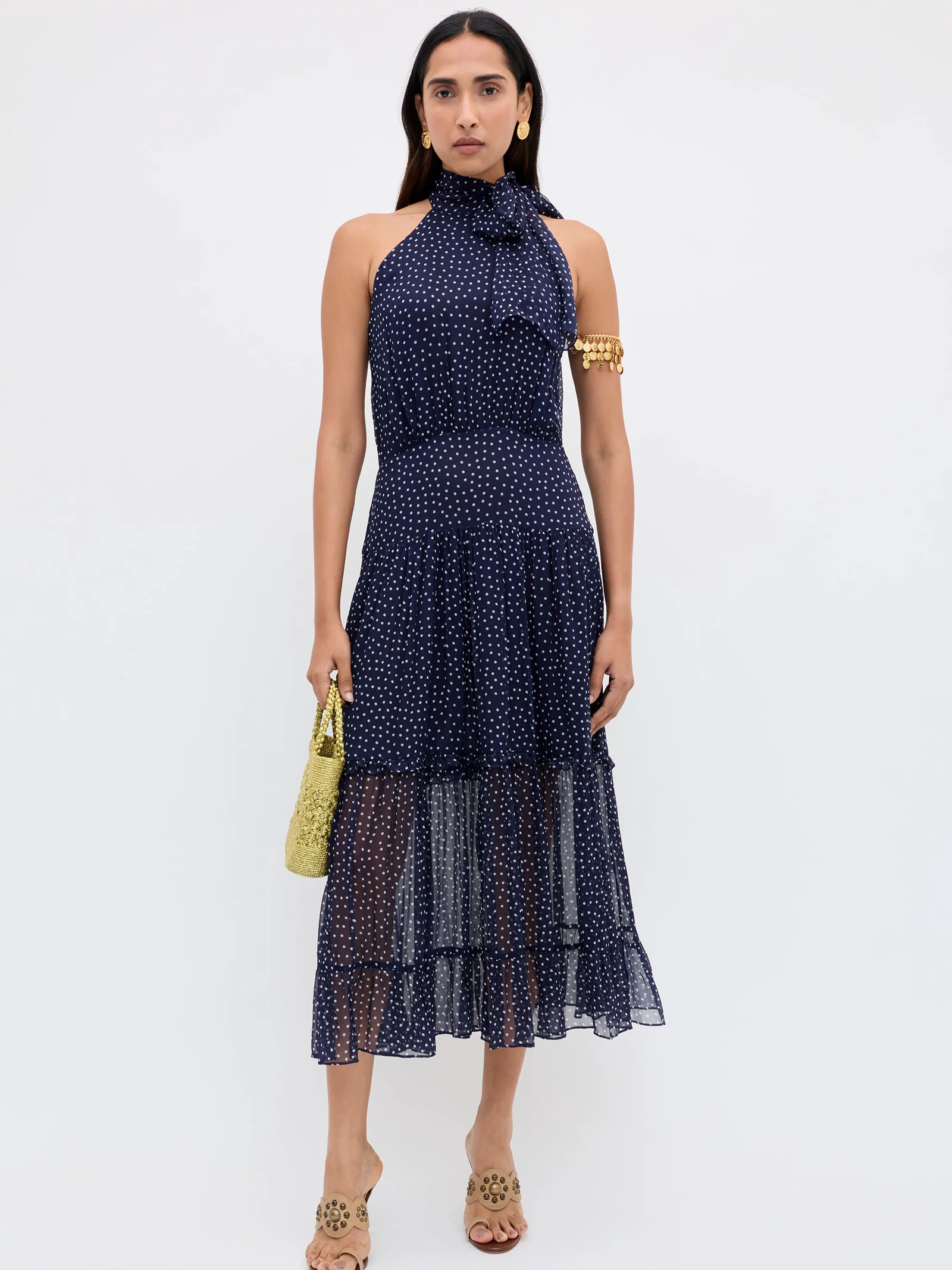 A woman stands against a white background, wearing the Abi - Mini Navy Polka Dot georgette halter-neck occasion dress with tan sandals, gold earrings, a gold arm cuff, and holding a textured yellow handbag.