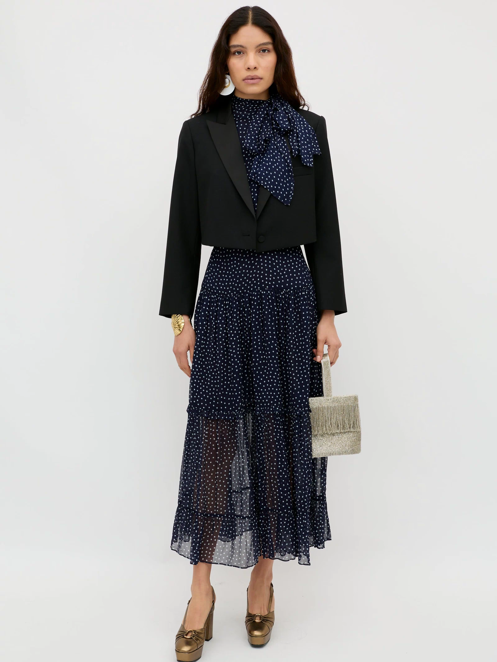 A woman stands against a plain white background wearing the Abi - Mini Navy Polka Dot dress, styled with gold heels and a small textured handbag—perfect as an occasion dress.