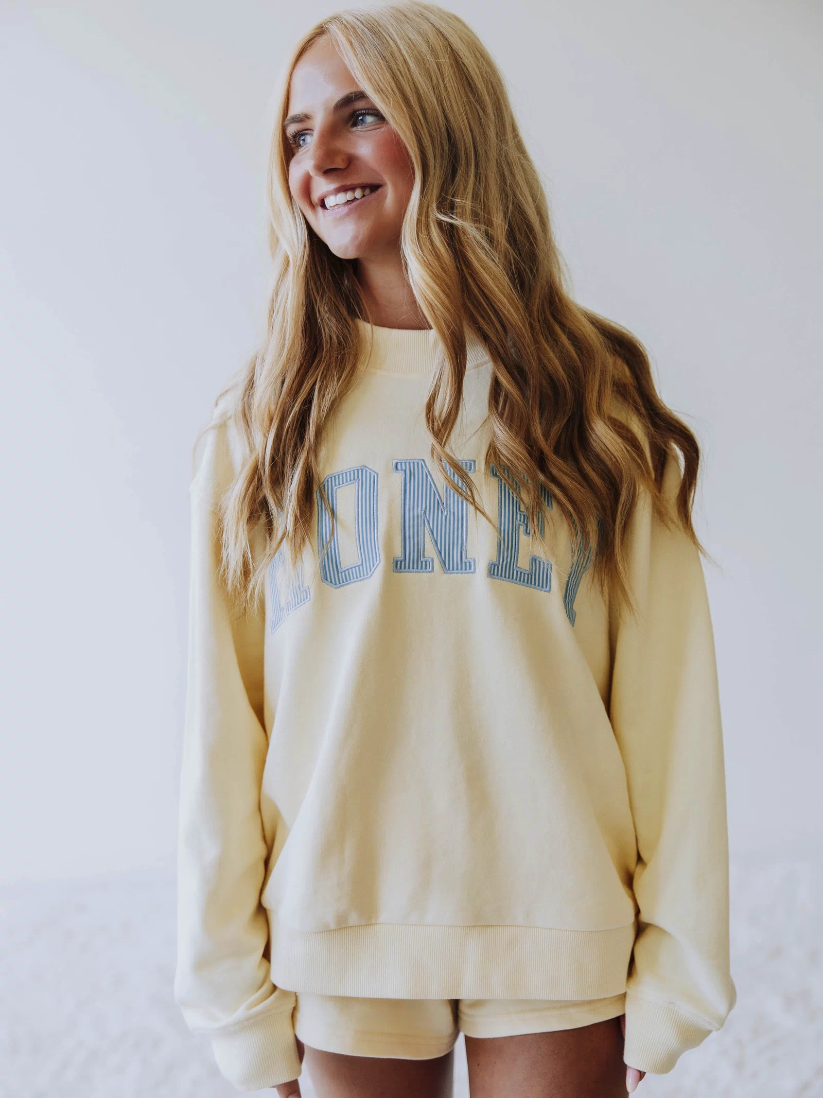Girl wearing a soft, yellow two piece set. Top says HONEY on the front. The HONEY has blue lines going through each letter. Shorts are same yellow as top. 