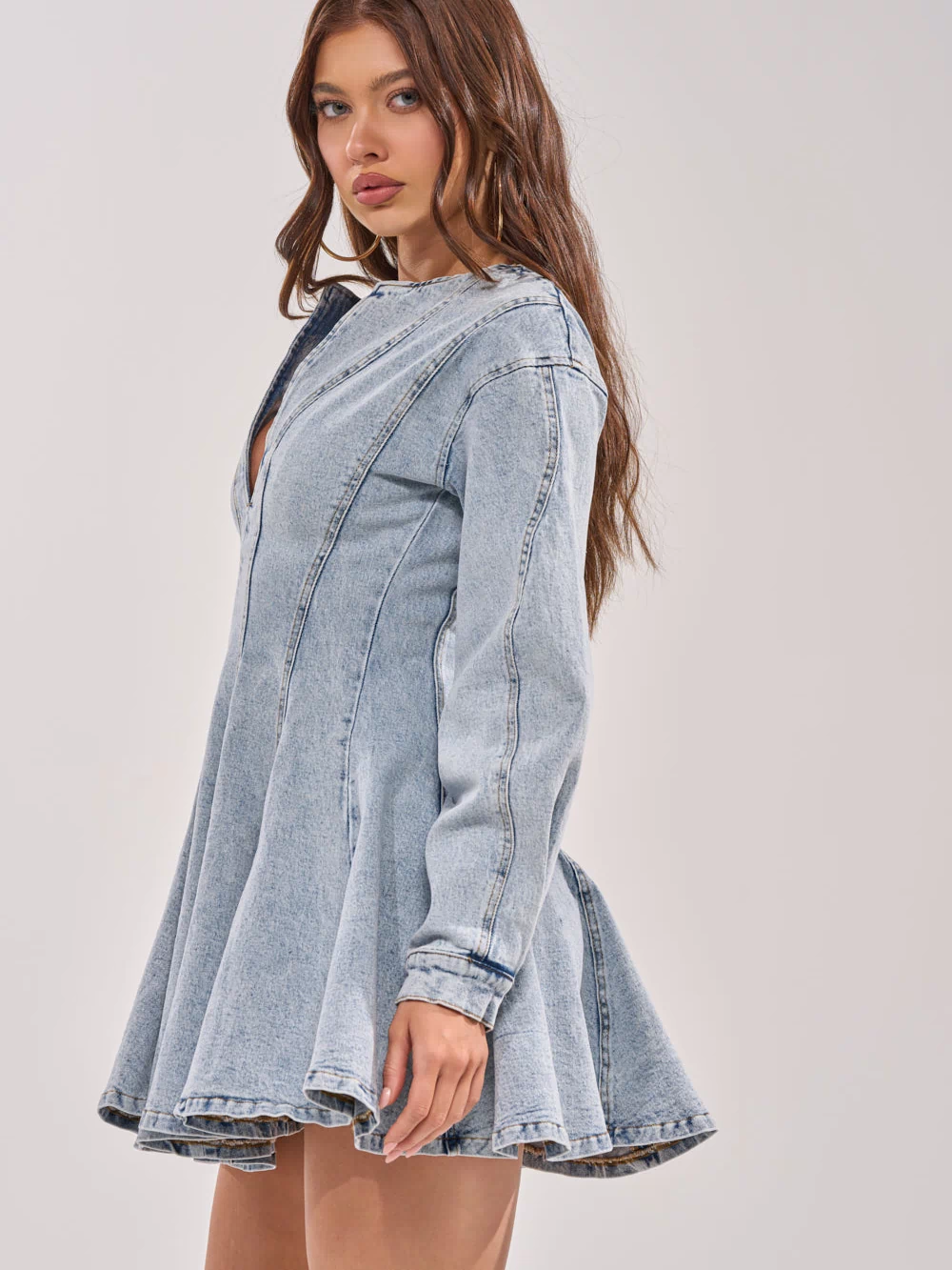 A woman with long brown hair models the ALL ABOUT MY BUSINESS DENIM MINI DRESS, a light wash fit and flare mini dress with balloon sleeves, standing against a plain light background and looking at the camera.