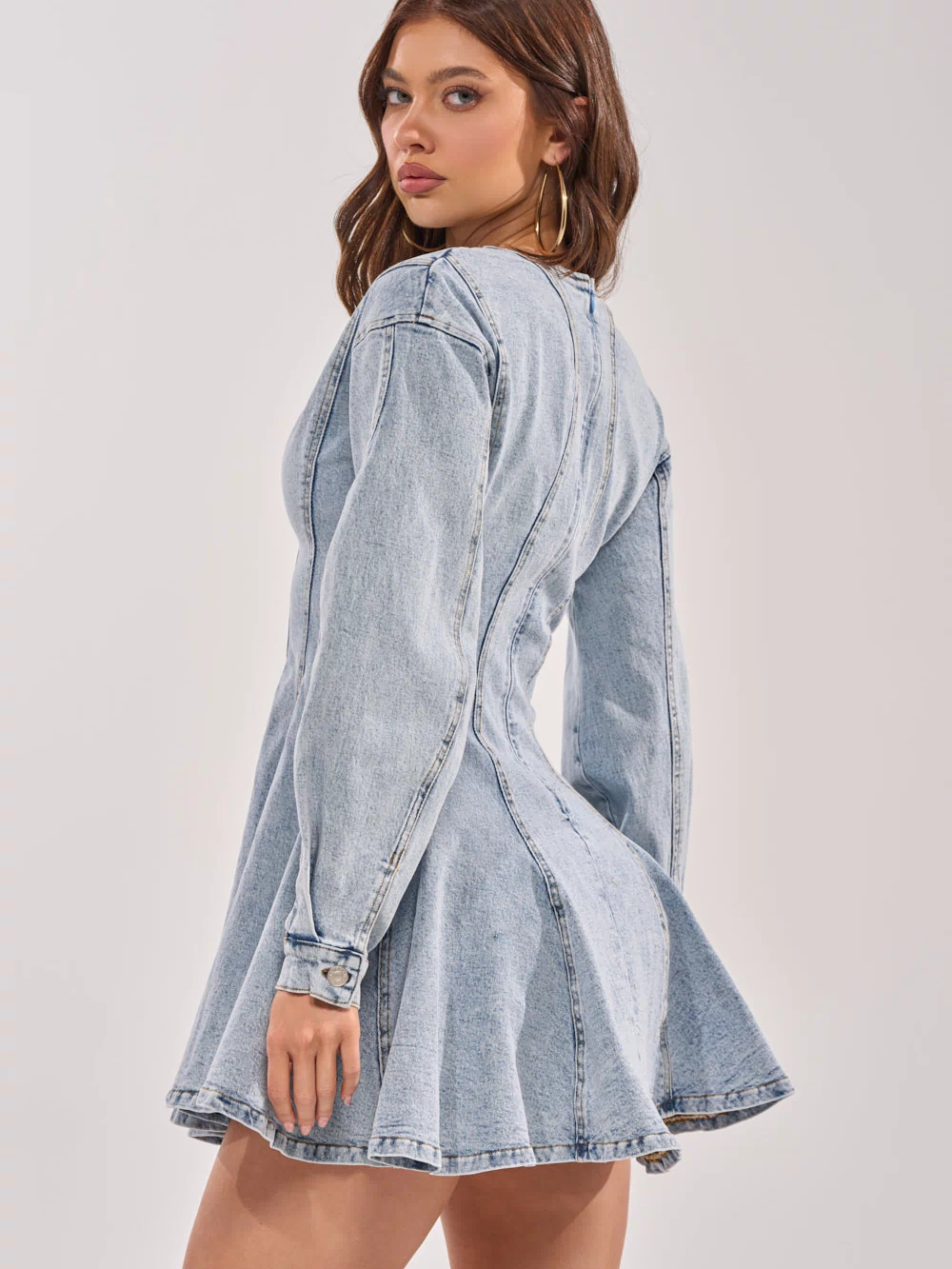 A woman with wavy brown hair models the ALL ABOUT MY BUSINESS DENIM MINI DRESS, featuring balloon sleeves and a fit-and-flare mini skirt. She stands sideways, glancing over her shoulder at the camera against a plain light background.