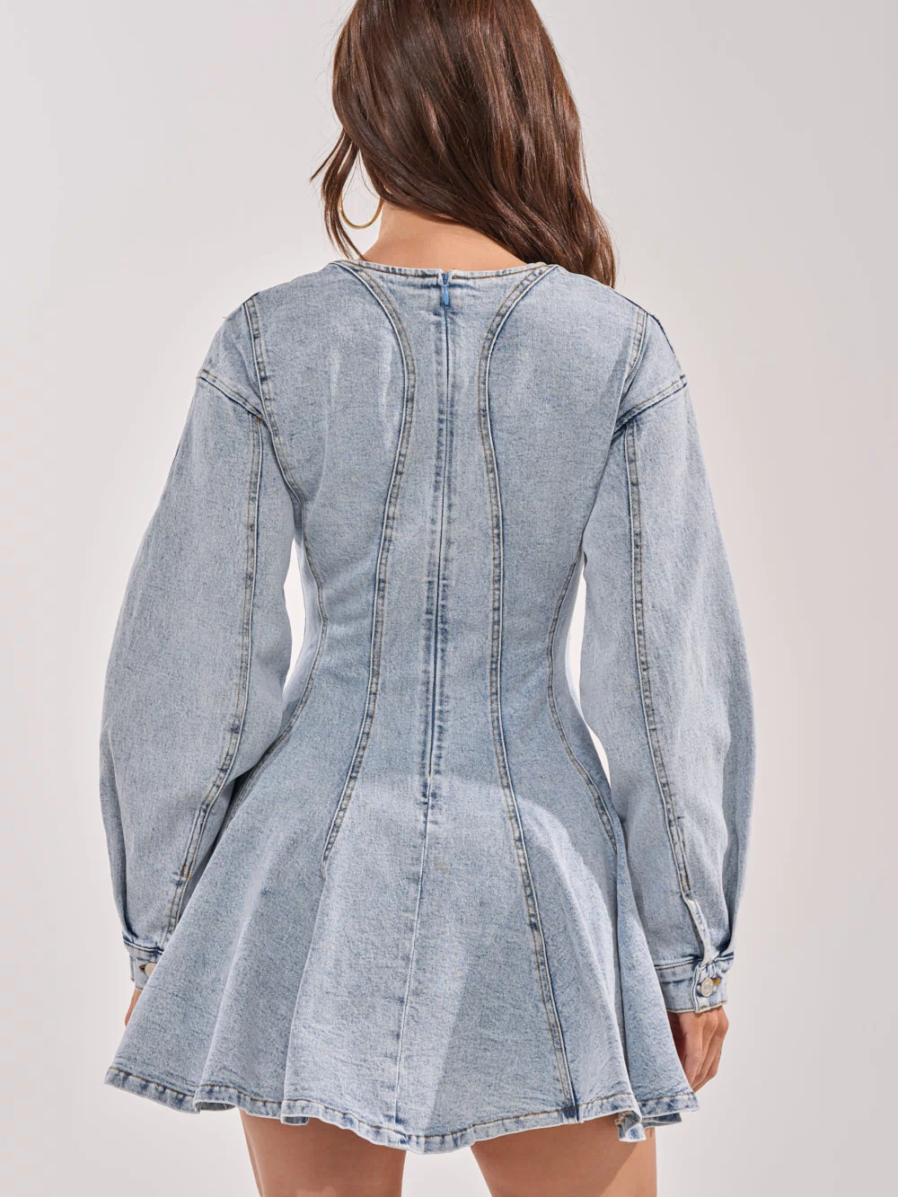 A woman stands with her back to the camera, wearing the ALL ABOUT MY BUSINESS DENIM MINI DRESS featuring a light wash, fitted waist, and flared skirt. Her long brown hair is down against a plain, light background.