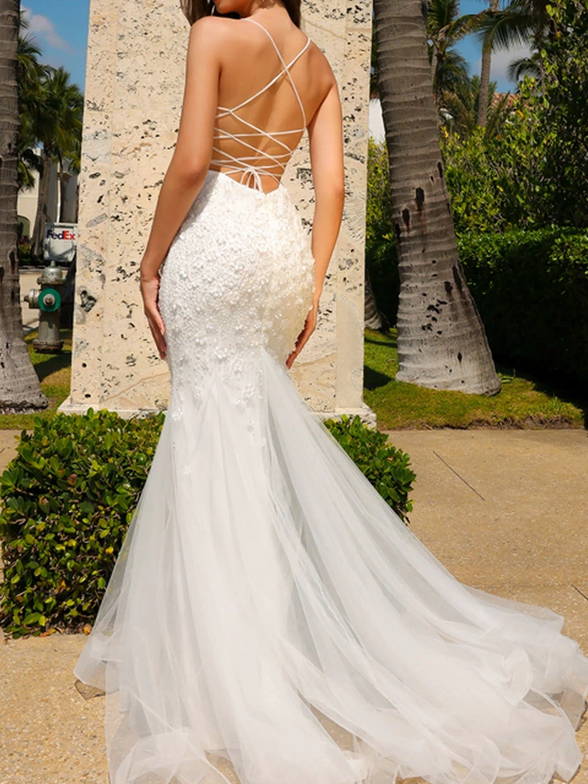 A woman stands outdoors near palm trees and a stone wall, wearing the Floral-Embellished Long Prom Dress with Train SU066, a white backless mermaid gown with lace-up straps and subtle floral details.