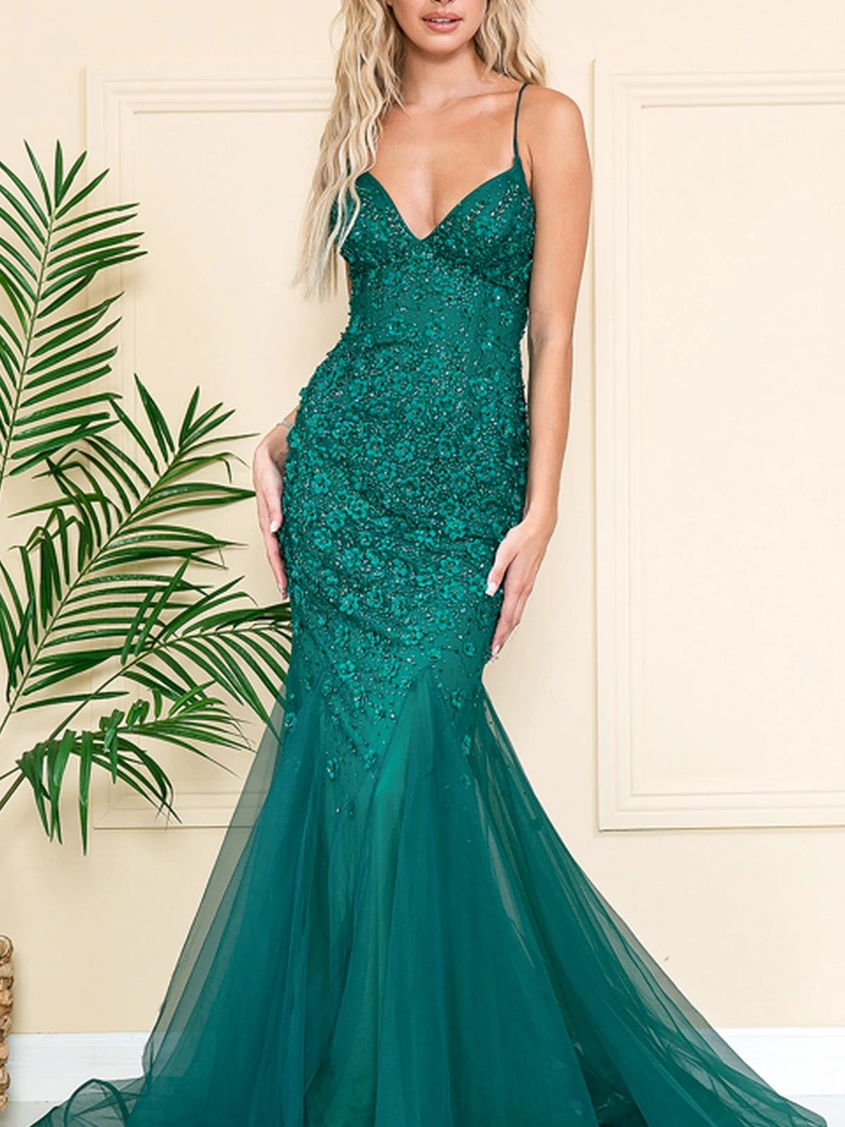 A woman in the Floral-Embellished Long Prom Dress with Train SU066, a fitted mermaid gown with thin straps and a flared tulle skirt, stands before a neutral wall and leafy plant.