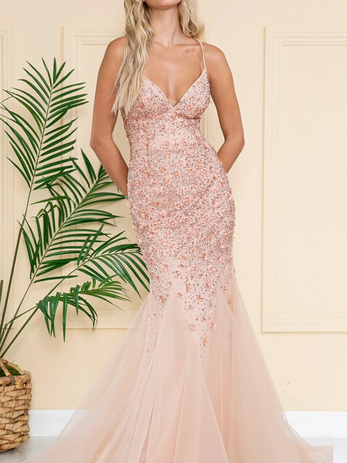A woman stands indoors in the Floral-Embellished Long Prom Dress with Train SU066—a fitted, blush mermaid gown with thin straps, floral details, and tulle skirt. A green potted plant sits beside her against a cream wall.