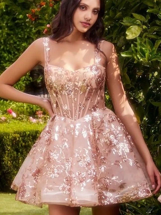 A woman stands outdoors in the Short Sequin-Print Babydoll Hoco Dress A1347, a sleeveless pink cocktail dress with a fitted bodice and shimmering detail. Greenery and flowers create a picturesque background.