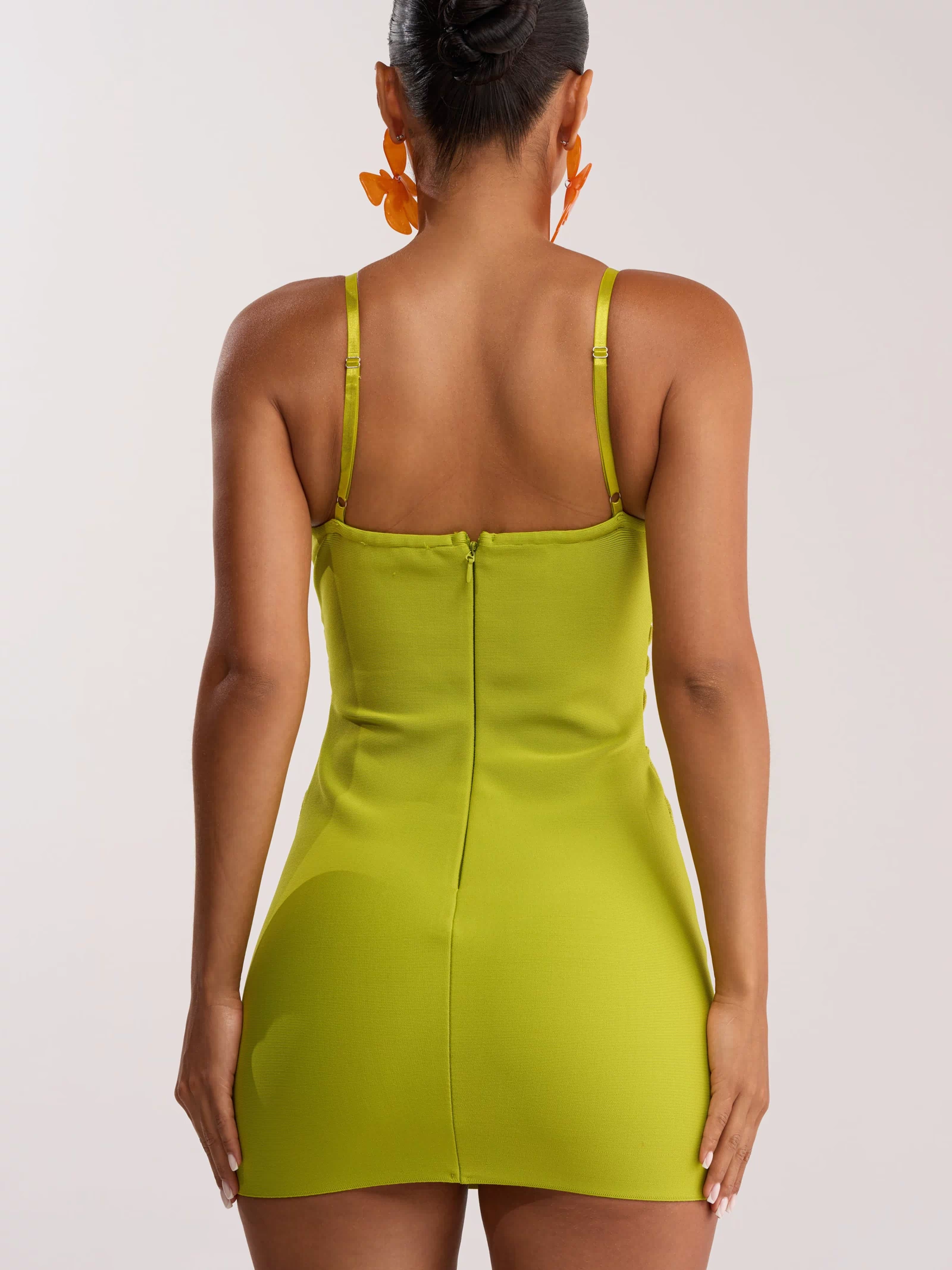 A woman with her hair in a bun wears the ANYTHING BUT BASIC MINI DRESS IN GREEN, a bright green bodycon mini dress with spaghetti straps and a visible back zipper, as she faces away from the camera.