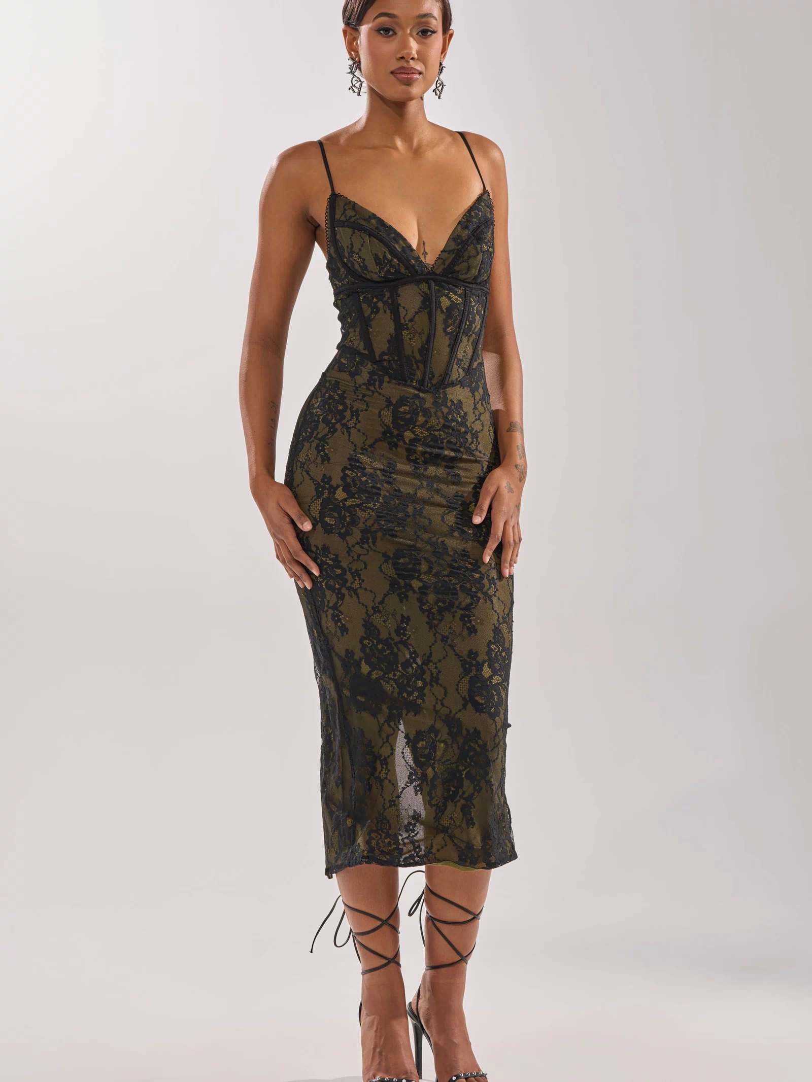 A woman poses against a light background in the BETTER BELIEVE IT LACE MAXI DRESS—a fitted, sleeveless black lace gown with thin straps and a deep neckline—styled with short hair, dangling earrings, and strappy high heels for an elegant evening look.