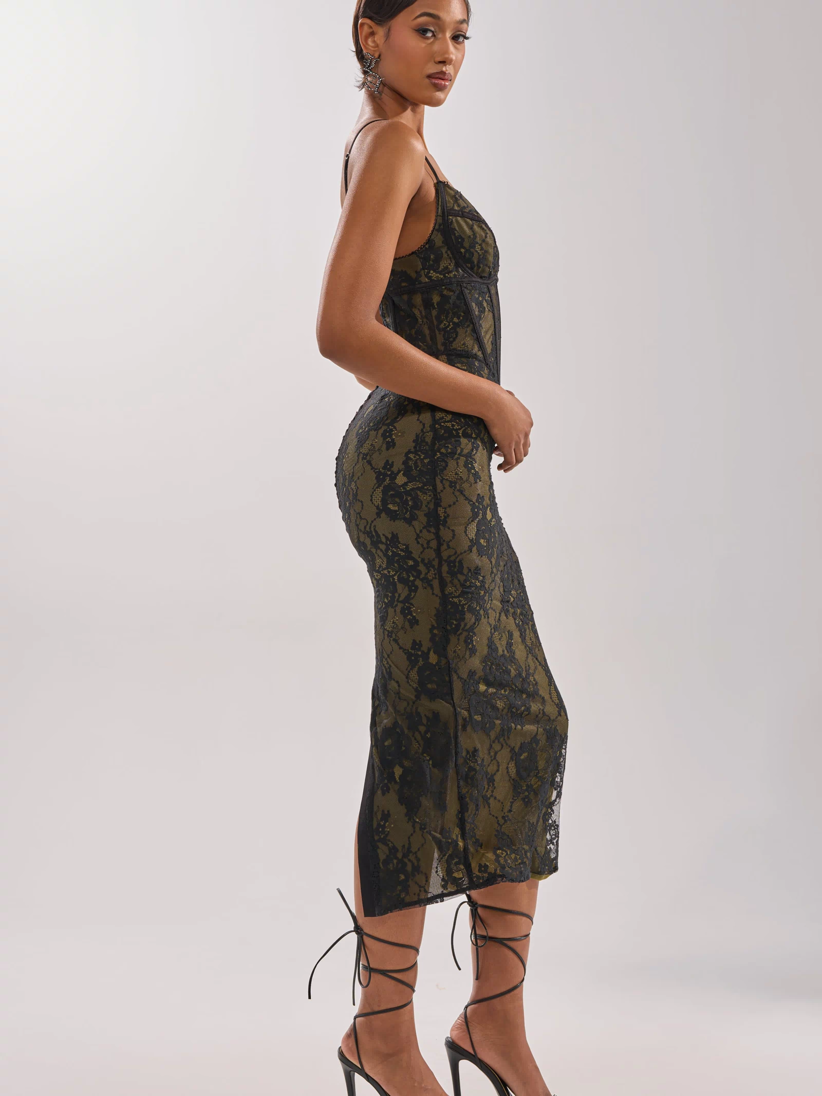 A woman stands sideways in the BETTER BELIEVE IT LACE MAXI DRESS—a sleeveless, form-fitting maxi dress with a corset bodice—paired with strappy black heels against a light neutral background.