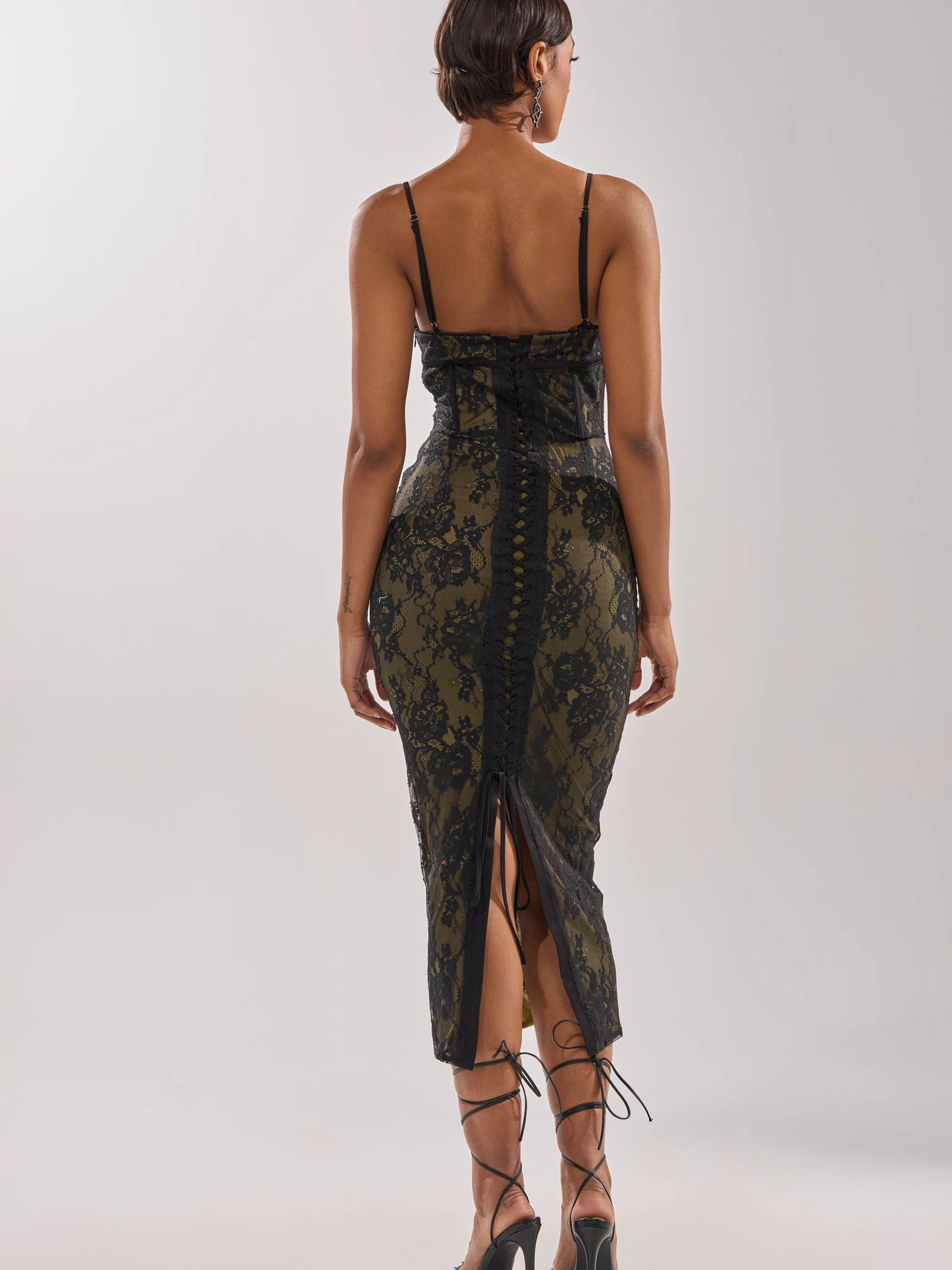 A person models the BETTER BELIEVE IT LACE MAXI DRESS, a form-fitting, dark green maxi with spaghetti straps, corset bodice, lace-up back, and intricate lace detailing, paired with high-heeled strappy sandals.