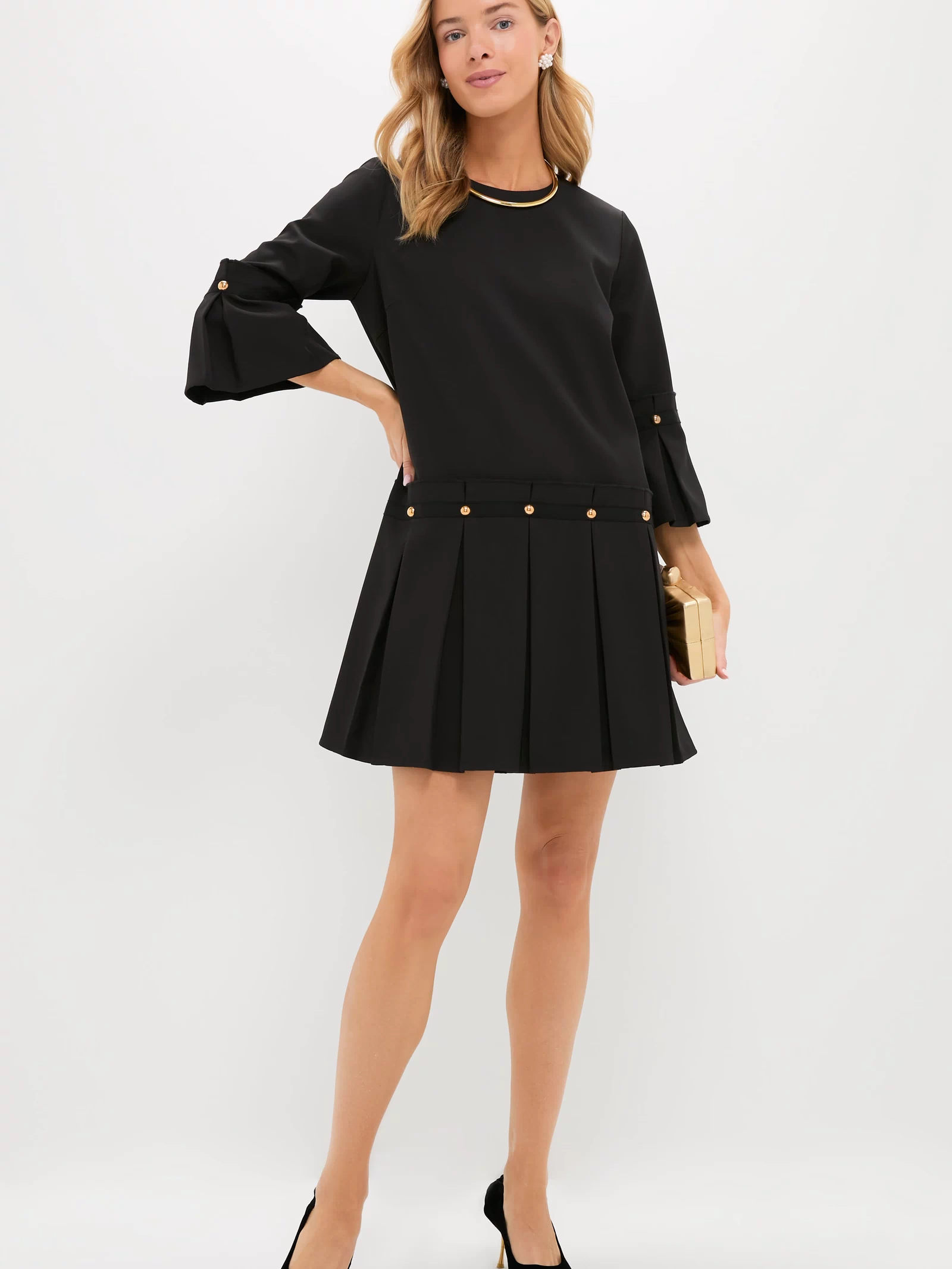 A woman stands against a white background wearing Tuckernuck's Kennedy Dress—a chic little black dress with a pleated skirt, gold button details, black heels, and a gold clutch.