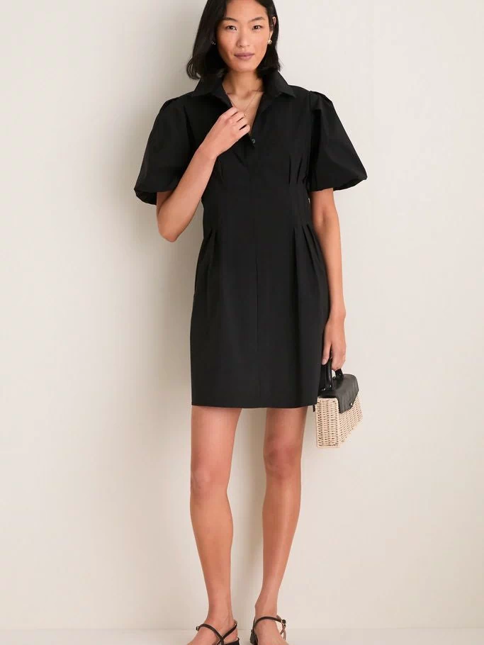 Woman models the Black Mini Delaney Dress, a classic, high-end shirt dress with puff sleeves and pleated waist, styled with black sandals and a woven handbag for an effortless, fun look.