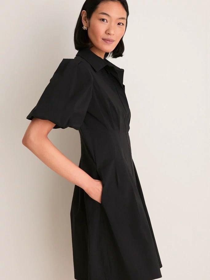Woman in classic Black Mini Delaney Dress with puff sleeves and pleated waist, embodying effortless, high-end style for versatile fun occasions, hands in side seam pockets.