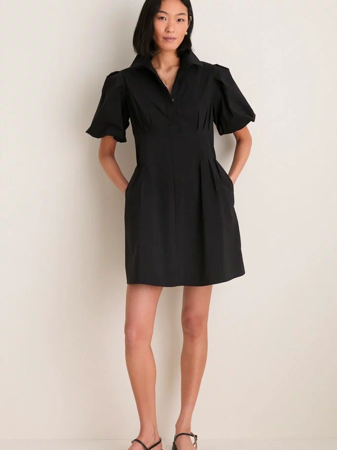 Woman in Black Mini Delaney Dress with puff sleeves and pleated waist, paired with strappy sandals, showcasing an effortless, classic look for versatile, high-end style.
