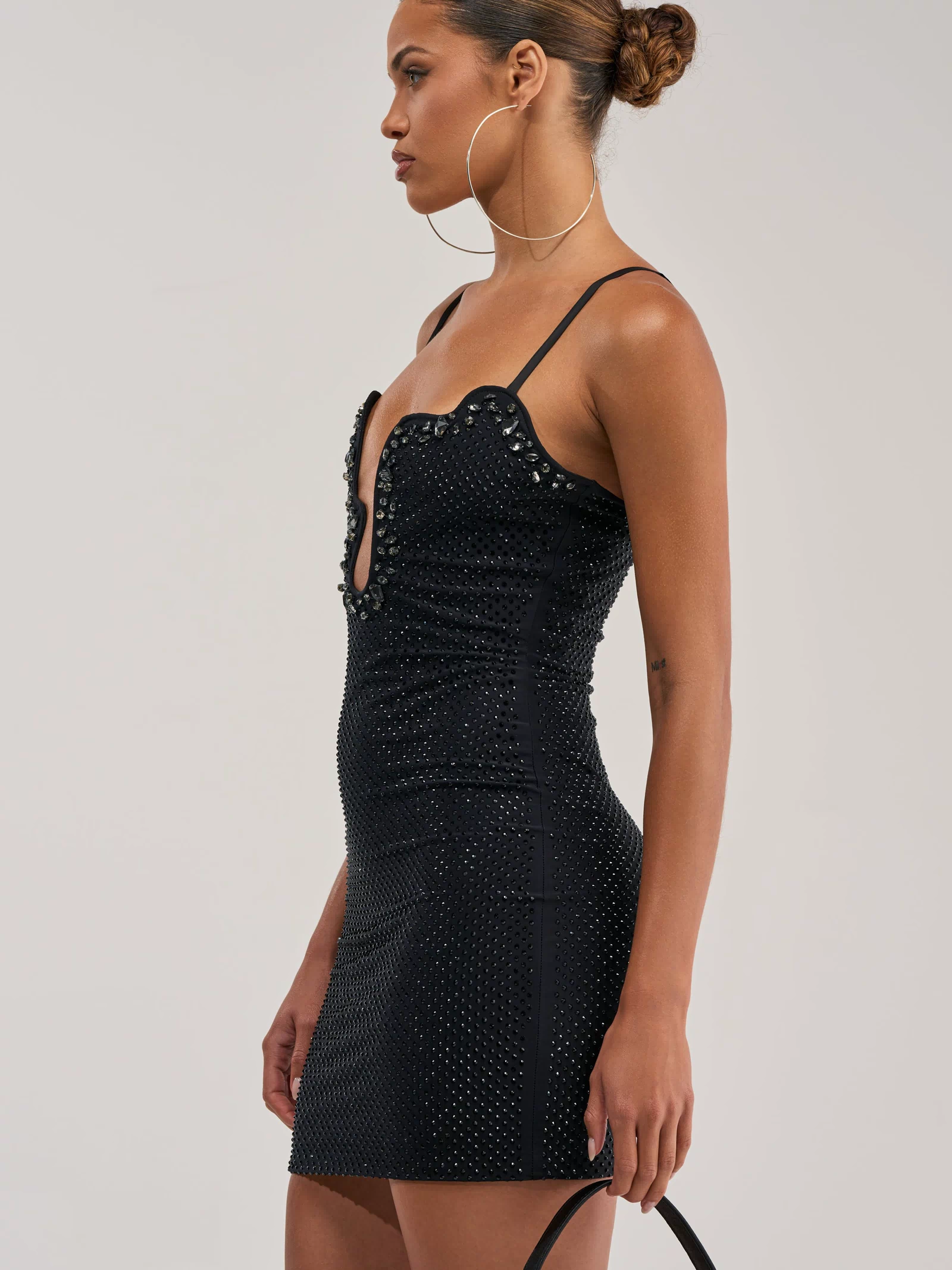 Wearing THE AFTERPARTY MINI DRESS—a fitted black rhinestone mini with a wavy neckline—a woman with her hair in a bun poses against a plain background, styled with large hoop earrings and a black handbag.