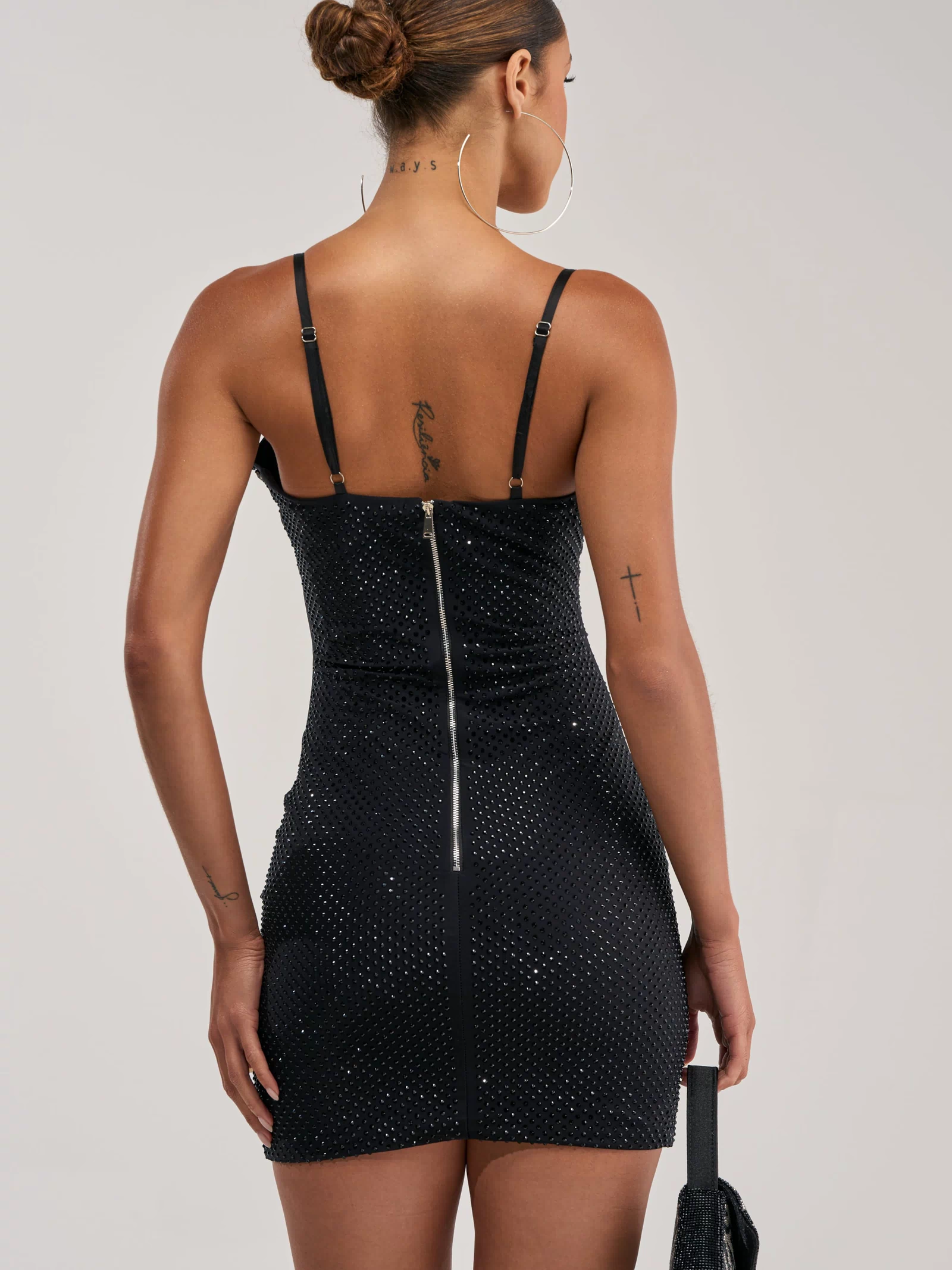 A woman with a neat bun is shown from the back wearing THE AFTERPARTY MINI DRESS, a black rhinestone mini dress with thin straps and a visible zipper. She holds a small black purse and has tattoos on her neck, arm, and back.