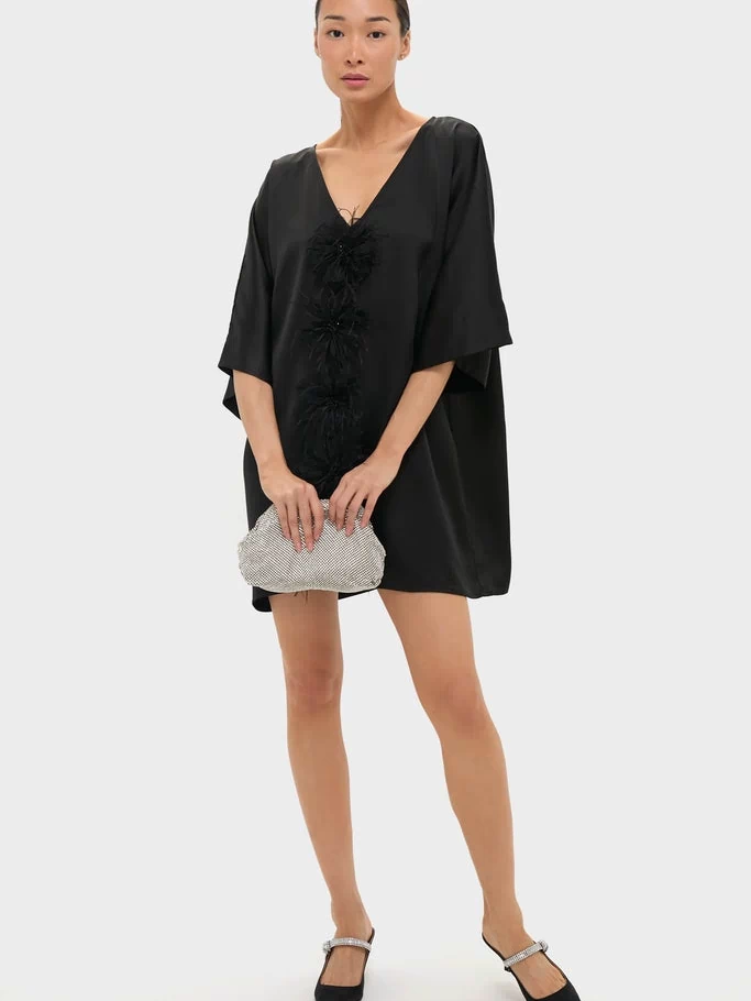 The Black Satin Diane Mini Dress features a classic V-neck, feathered rosettes, and kimono sleeves, paired with a silver clutch and sparkly slingback flats for an effortless, fun, high-end look.