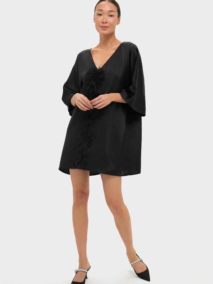 Woman models the classic Black Satin Diane Mini Dress, featuring a deep V-neck, kimono sleeves, and feather floral embellishments, embodying effortless elegance and high-end style.