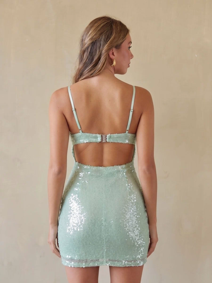 A woman shown from the back wearing a mint green sequined mini dress with thin straps and an open back, standing against a plain beige wall.