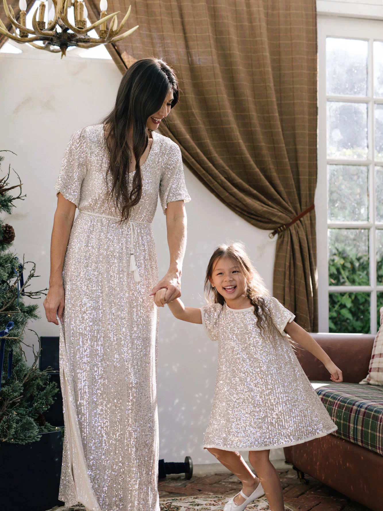 A long, shimmering silver sequin dress with a high neck, short sleeves, and a flowing maxi-length skirt, paired with a sequin-embellished child's dress in a matching style.
