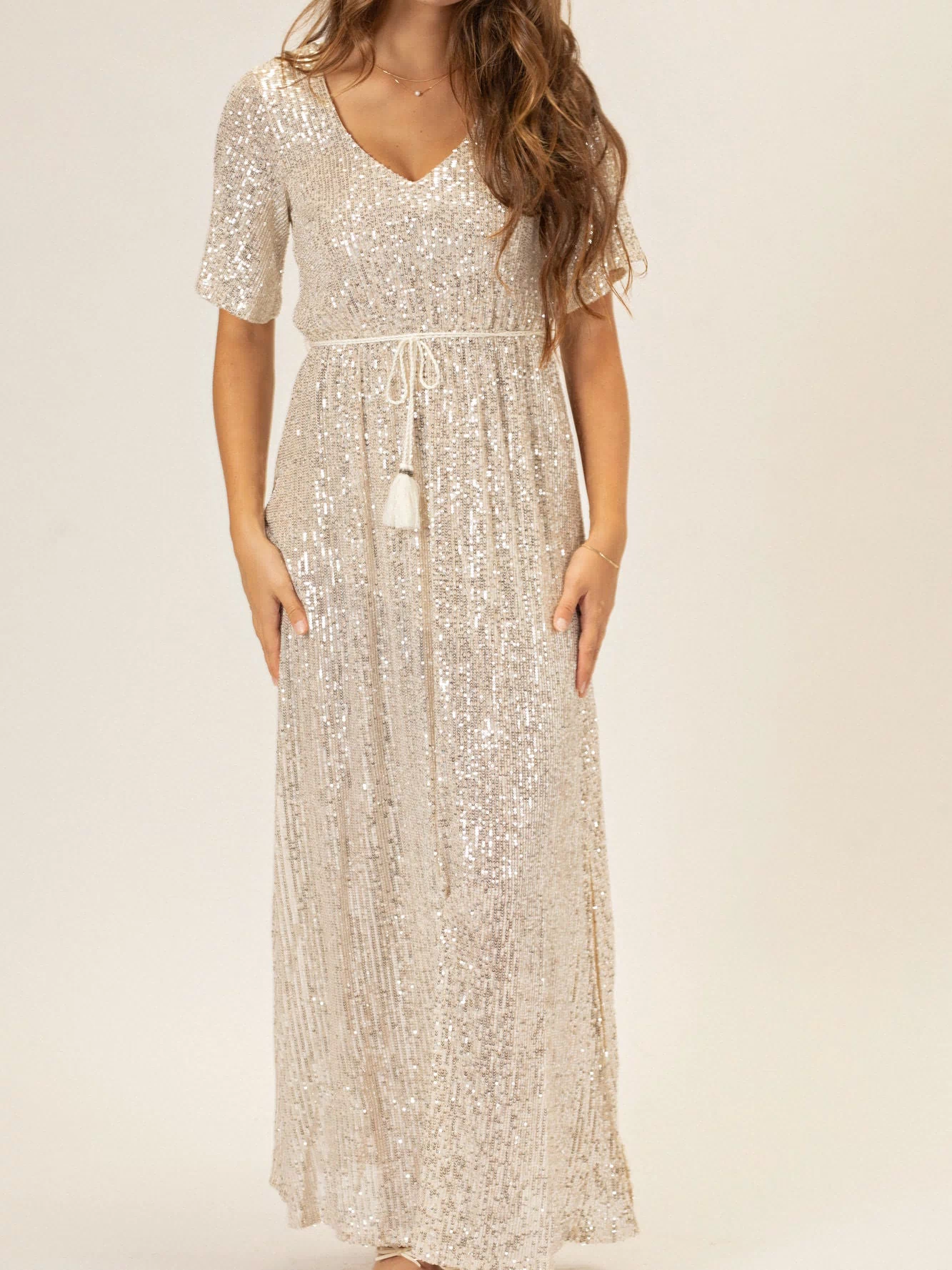 Glimmering silver sequin dress with a deep V-neckline, flared skirt, and tie-waist detail creating a flowing, elegant silhouette.