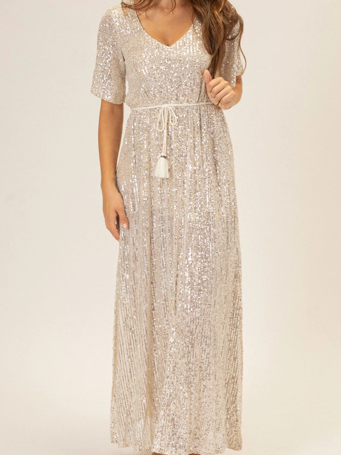 A long, sleeveless dress in a shimmering silver sequin fabric, with a V-neckline, a belted waistline, and a flowing maxi-length skirt.