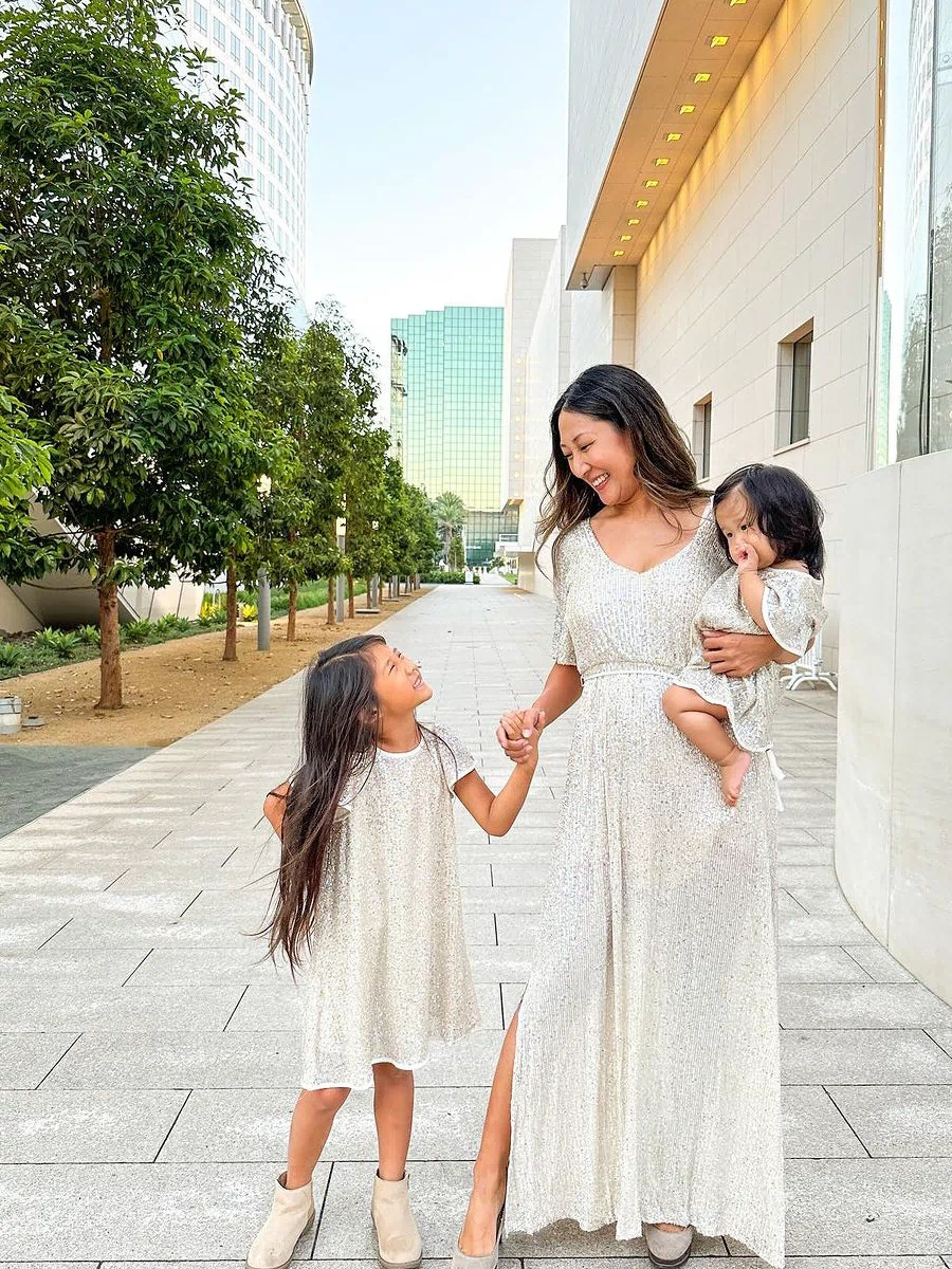 A woman in a long, sequined white dress stands with her two young daughters, all smiling and holding hands in a city setting with tall buildings in the background.