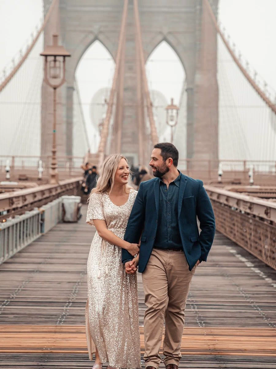 A woman in a sparkling silver sequin maxi dress stands hand-in-hand with a man in a navy suit on the Brooklyn Bridge, with the iconic bridge and cityscape in the background.