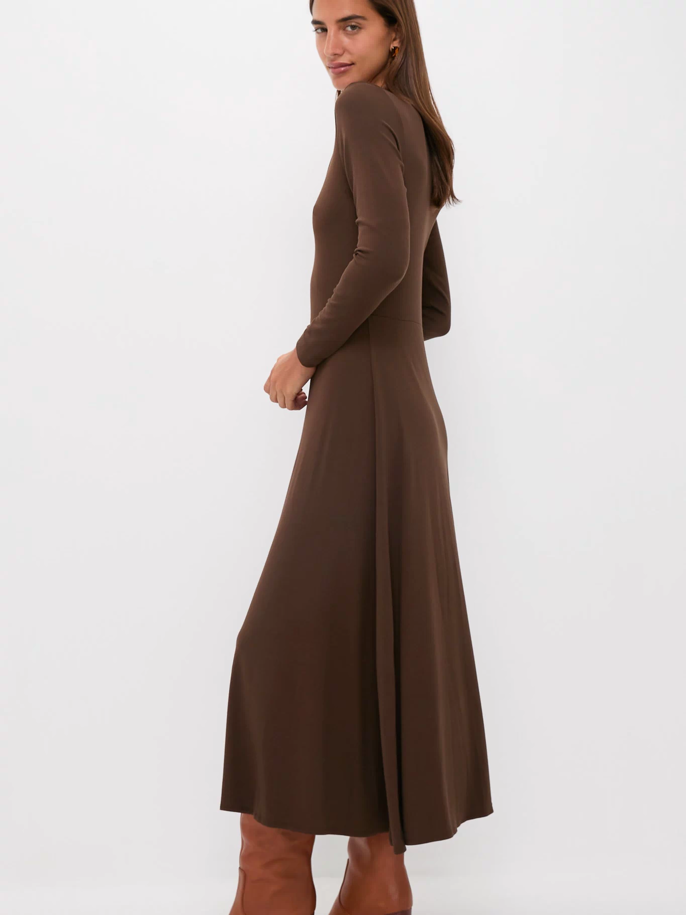 A woman stands sideways in a Tuckernuck Brown Sara Maxi knit dress with long sleeves and an ankle-length fit & flare silhouette, paired with brown heeled boots, against a plain white background.