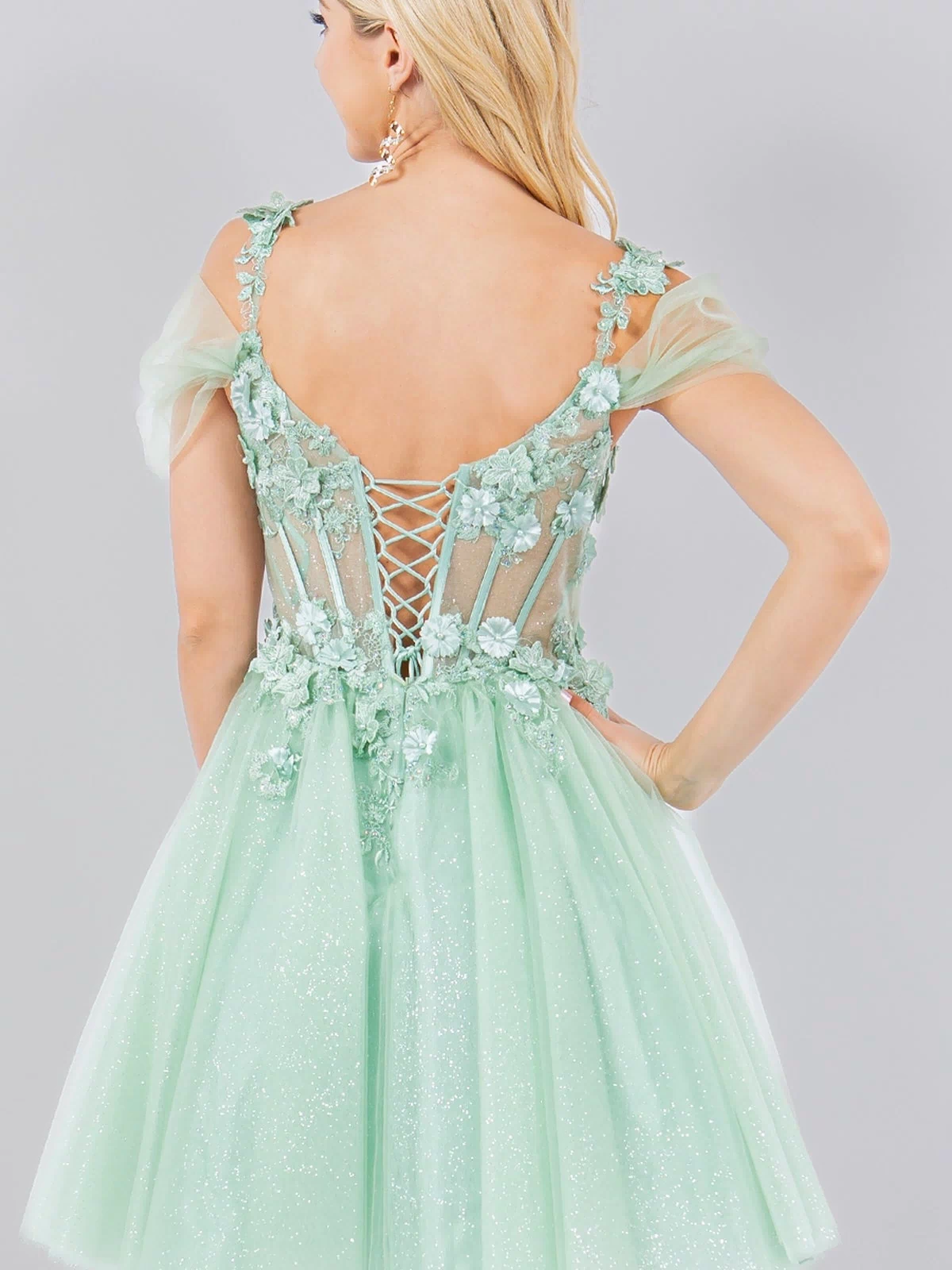 A woman in the Embellished Short Glitter-Tulle Homecoming Dress, featuring light green floral accents and a lace-up back, stands with her back to the camera.