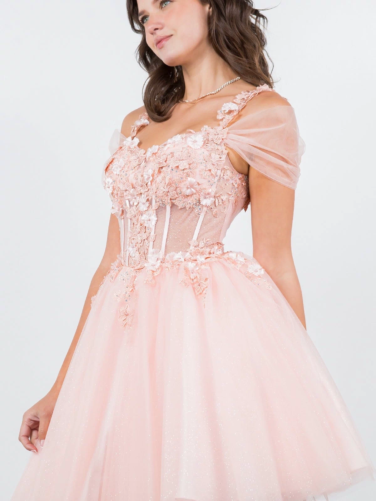 A woman poses against a plain background in the Embellished Short Glitter-Tulle Homecoming Dress, featuring light pink color, floral accents, sheer off-shoulder sleeves, and a short, sparkling tulle skirt.