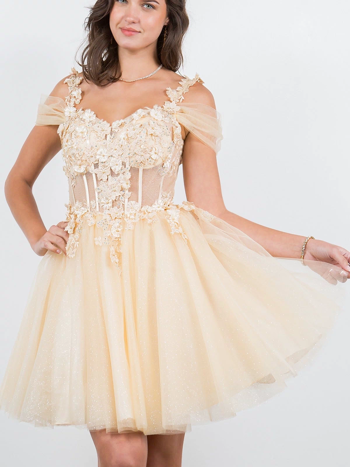 A young woman poses against a plain white background wearing the Embellished Short Glitter-Tulle Homecoming Dress, featuring an off-the-shoulder neckline, fitted bodice, and floral appliqué in light yellow.