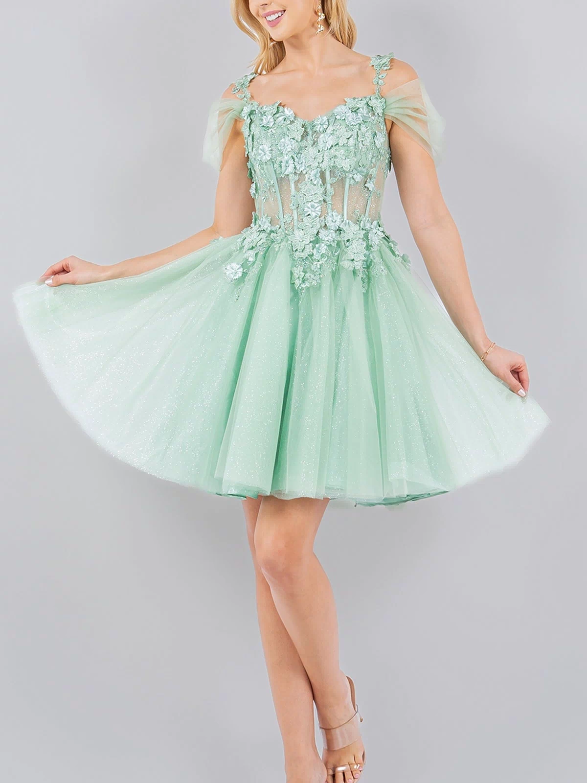Woman modeling the Embellished Short Glitter-Tulle Homecoming Dress, a light green off-shoulder style with sheer floral details, against a plain light gray background.