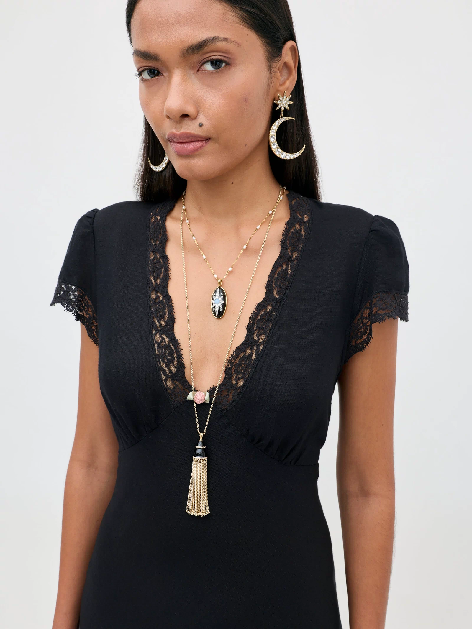 A woman with long dark hair wears the Clarice - Black vintage nightdress with lace trim, layered necklaces, and gold crescent moon earrings, standing confidently against a plain light background and looking at the camera.