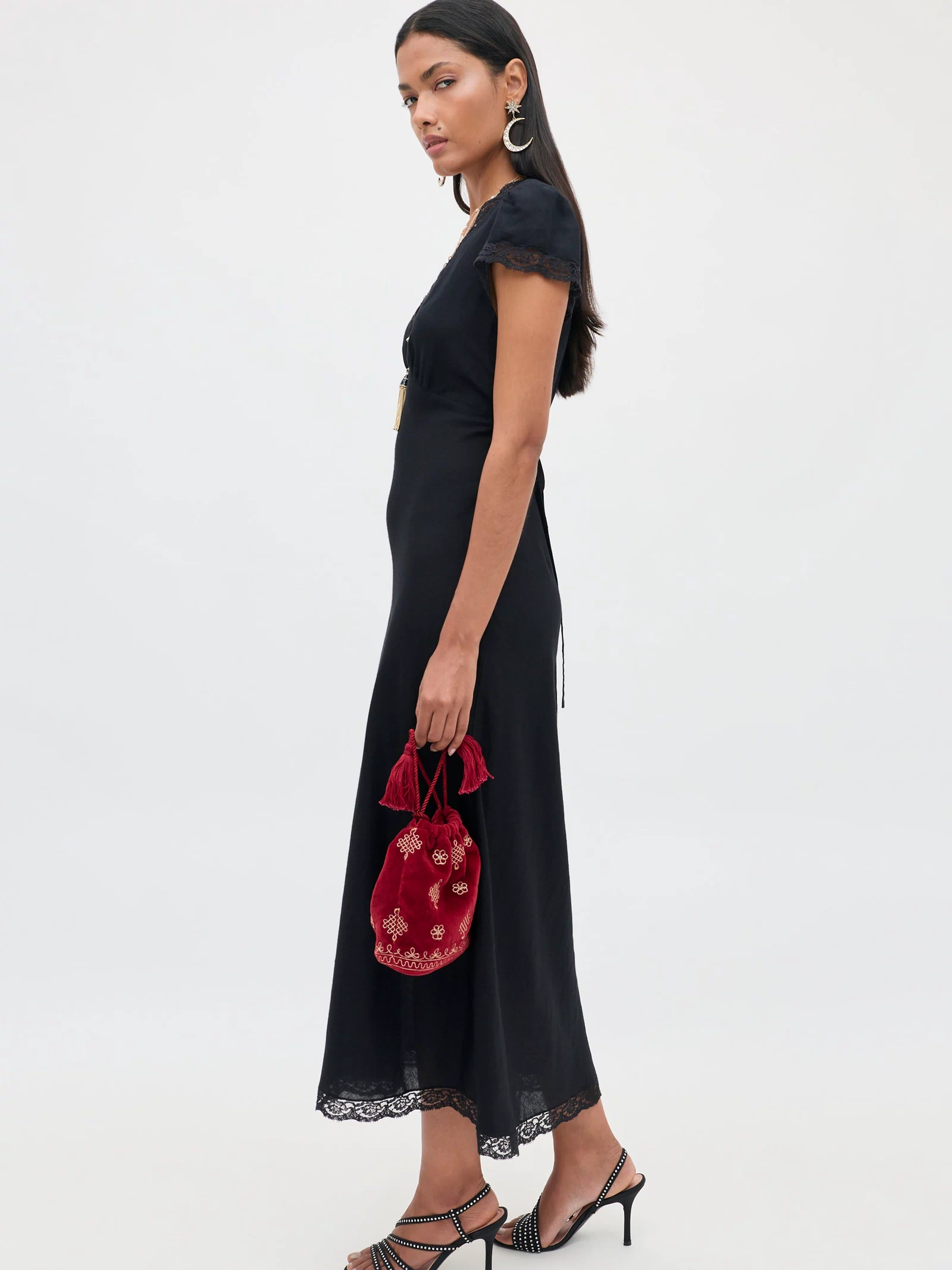A woman wearing the Clarice - Black midi dress with lace trim holds a small red embroidered bag, paired with black strappy heels and silver earrings, standing against a plain white background, evoking the elegance of a vintage nightdress.