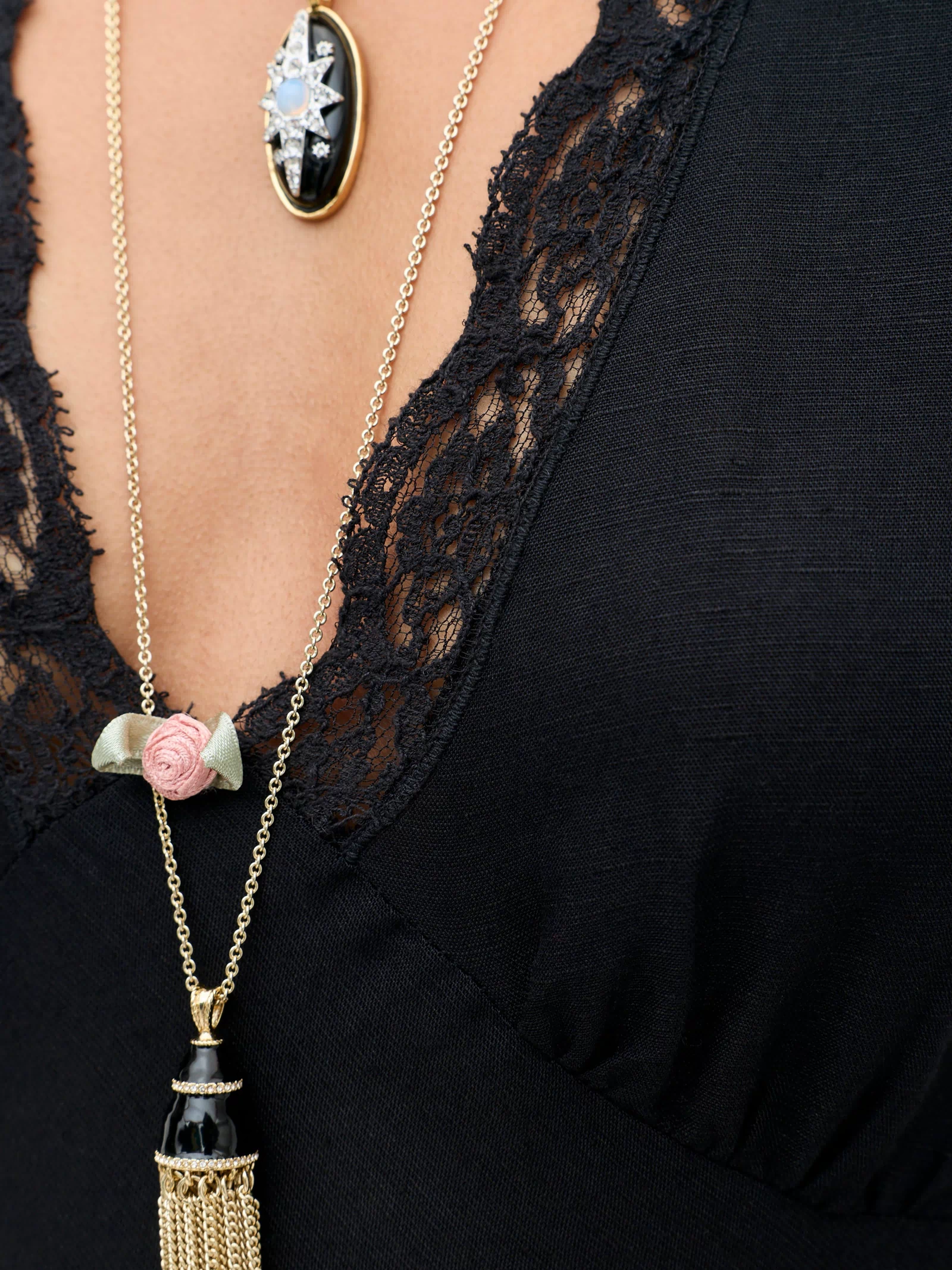 A close-up of a person wearing the Clarice - Black top, featuring vintage-inspired lace trim, along with two necklaces: one adorned with a black oval pendant and star, and the other with a small pink rose and gold tassel.