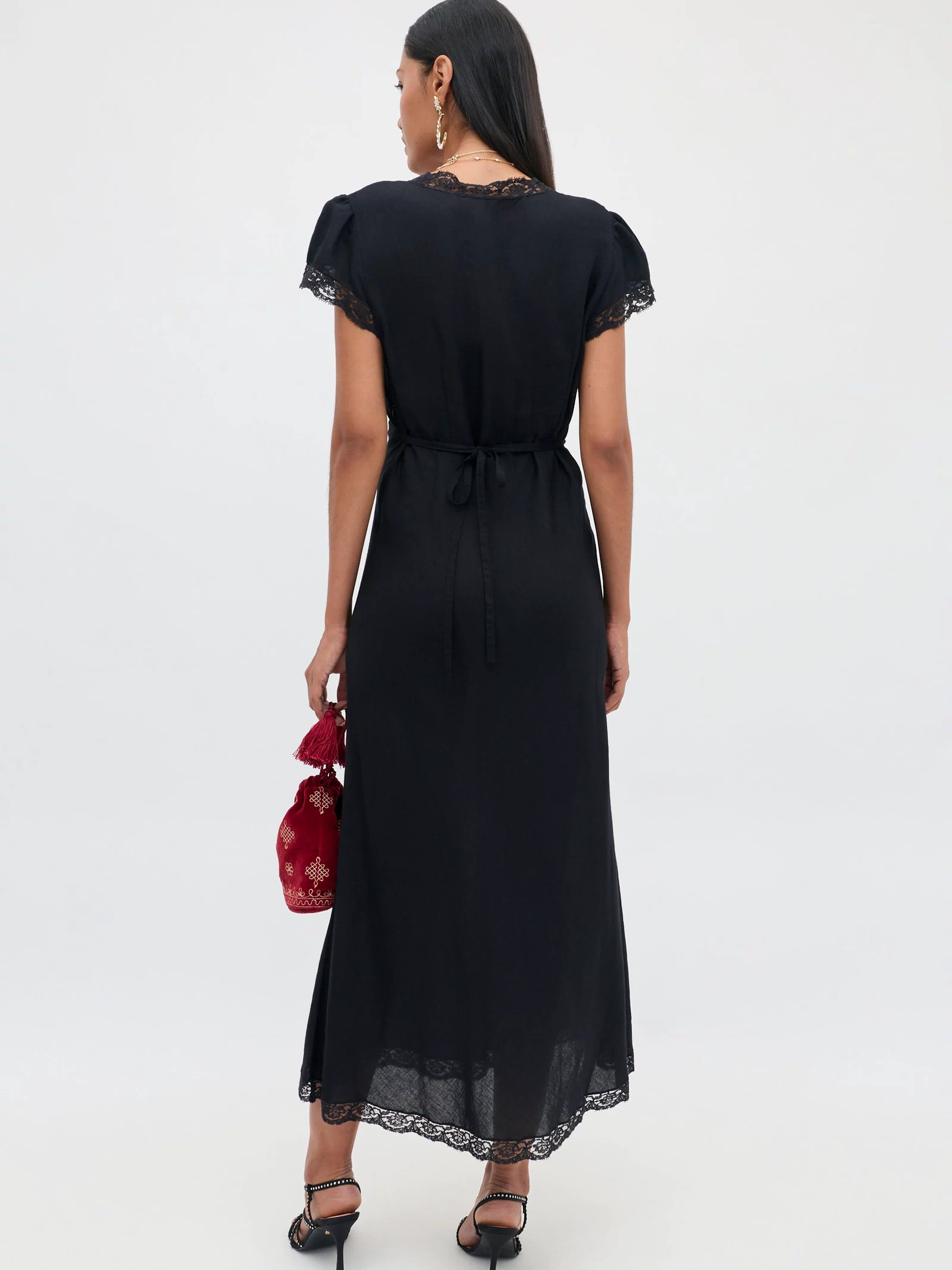 A woman wearing the Clarice - Black dress, featuring short sleeves and lace trim in a vintage-inspired nightdress style, holds a small red bag and stands in black heels with her back to the camera against a simple light background.