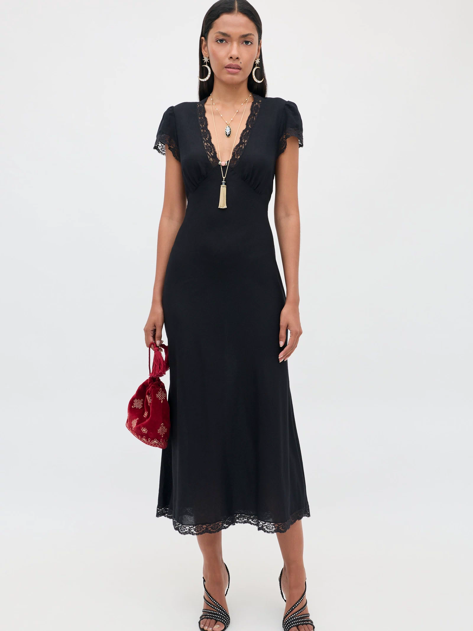 Wearing the Clarice - Black dress, a woman poses against a plain light background. The vintage nightdress-inspired midi features short sleeves and delicate lace trim. She accessorizes with gold necklaces, hoop earrings, black strappy heels, and holds a red velvet bag.