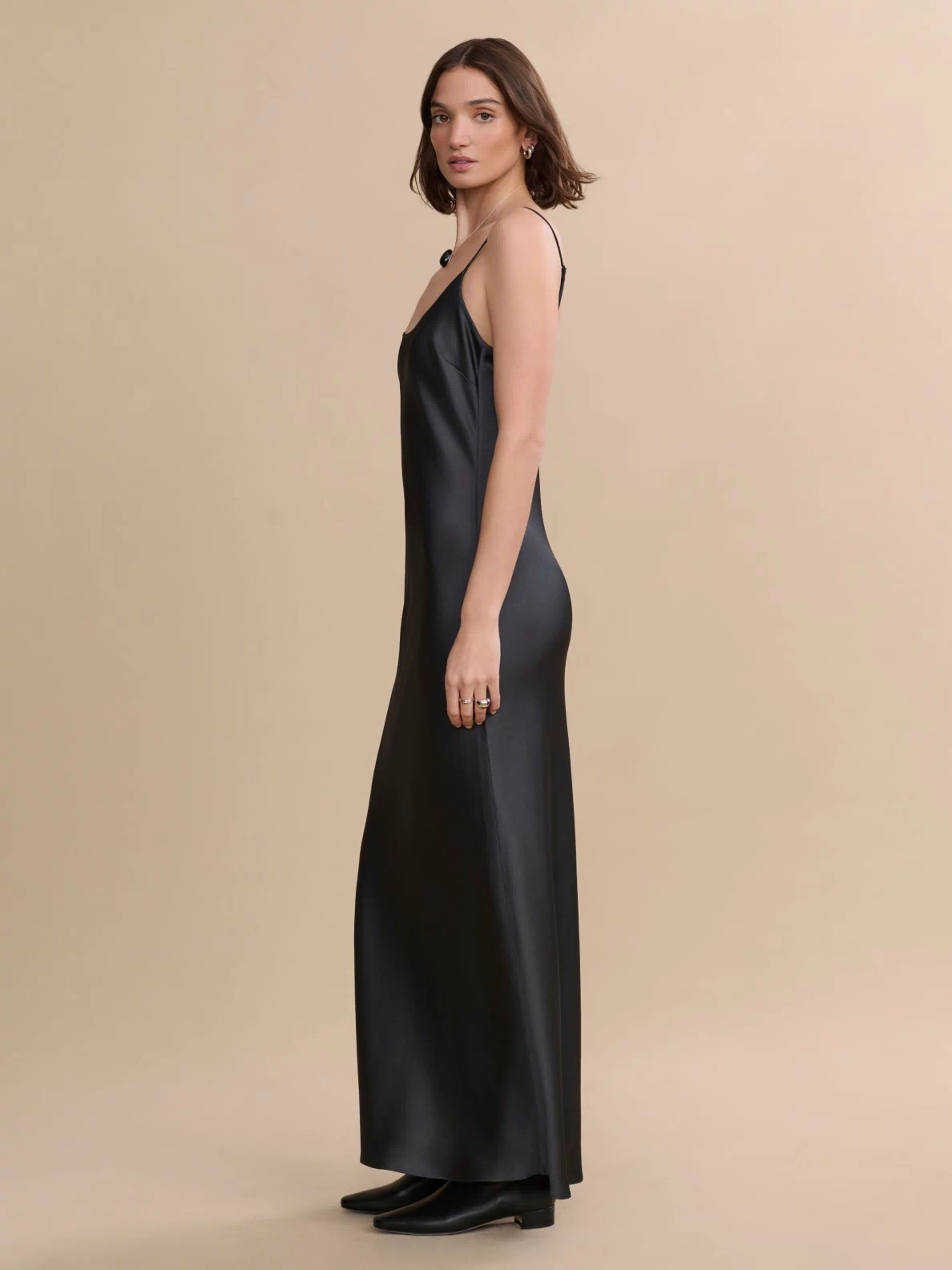 A woman stands sideways in the Cleo Slip Dress—long, black, with thin straps—paired with black boots against a beige background.