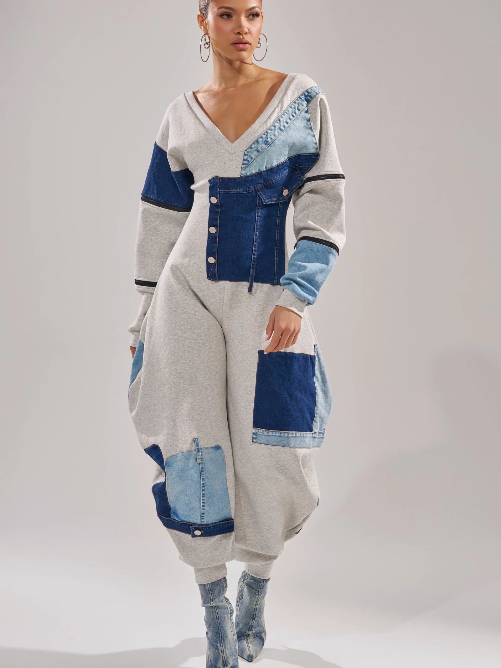 A woman wears the COMFY COZY PATCHWORK JUMPSUIT IN HEATHER GREY, blending grey sweatshirt fabric with blue panels. She styles this streetwear piece with blue textured heeled boots and large hoop earrings.