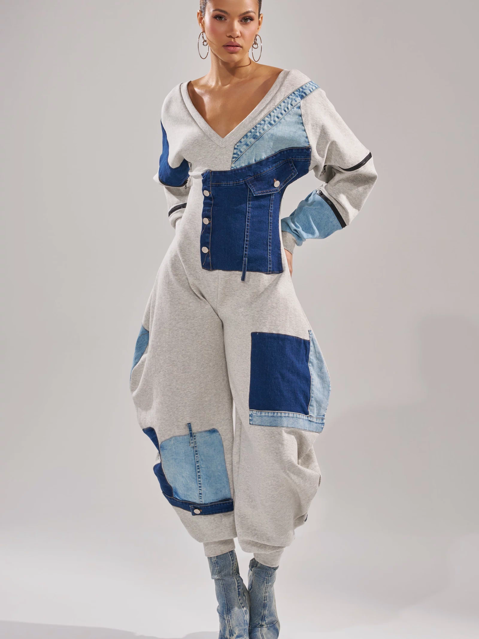 A woman models the COMFY COZY PATCHWORK JUMPSUIT IN HEATHER GREY, featuring patchwork denim and grey accents. She pairs it with matching denim boots and hoop earrings, posing confidently against a plain background.