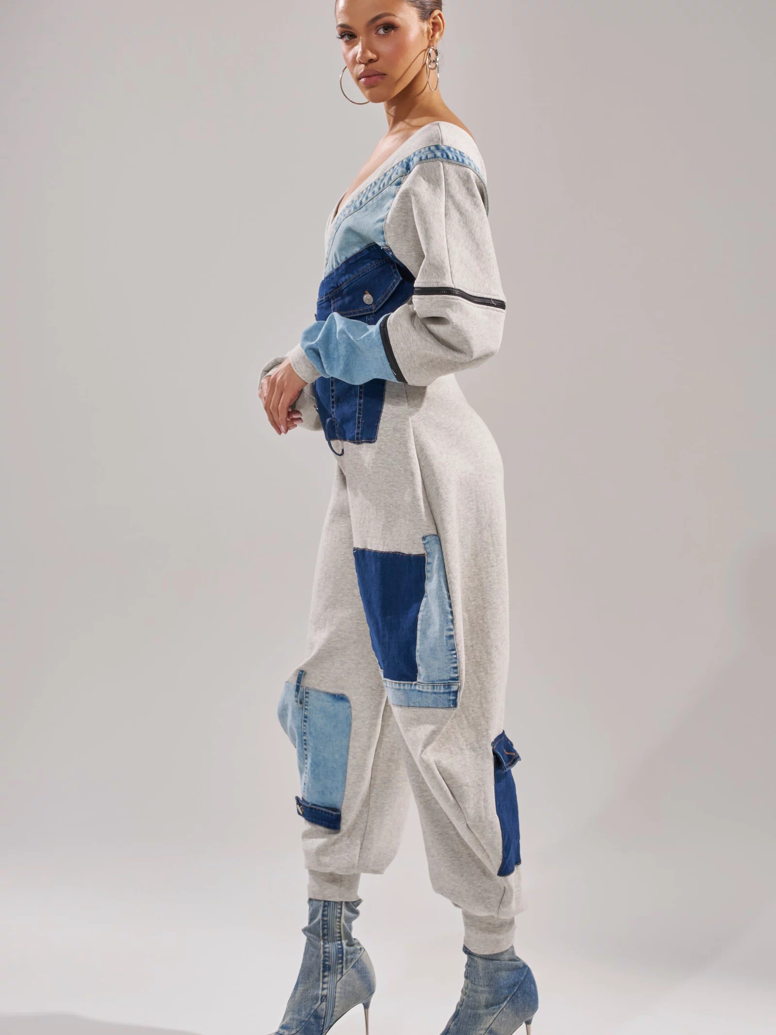 A woman models the COMFY COZY PATCHWORK JUMPSUIT IN HEATHER GREY, styled with hoop earrings and high-heeled denim boots, showcasing a chic one-piece streetwear look against a light-gray background.