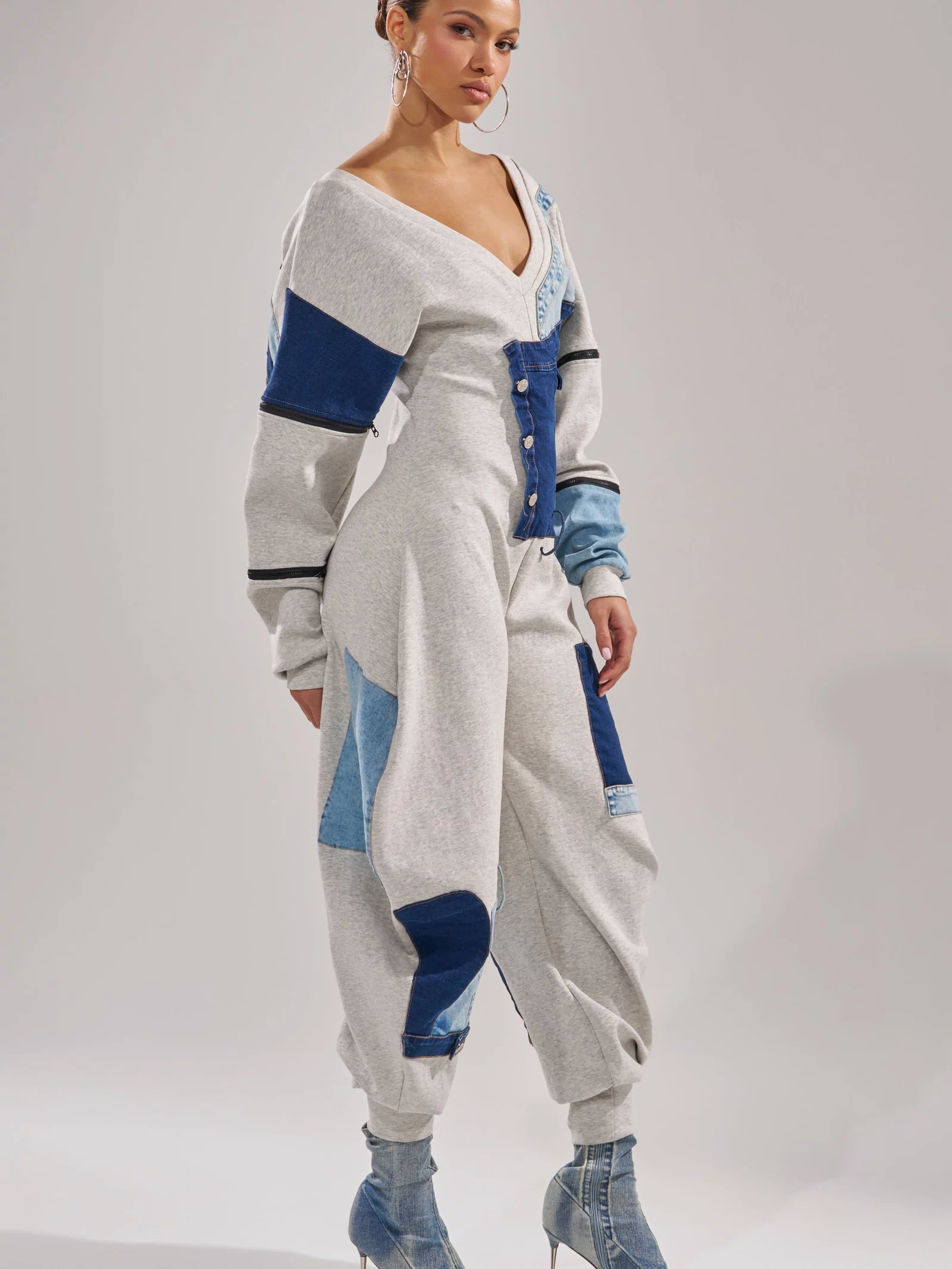 A woman radiates confidence in the COMFY COZY PATCHWORK JUMPSUIT IN HEATHER GREY, featuring blue denim patches, long sleeves, buttons, and matching denim ankle boots. She completes her chic streetwear look with hoop earrings and a neat bun.