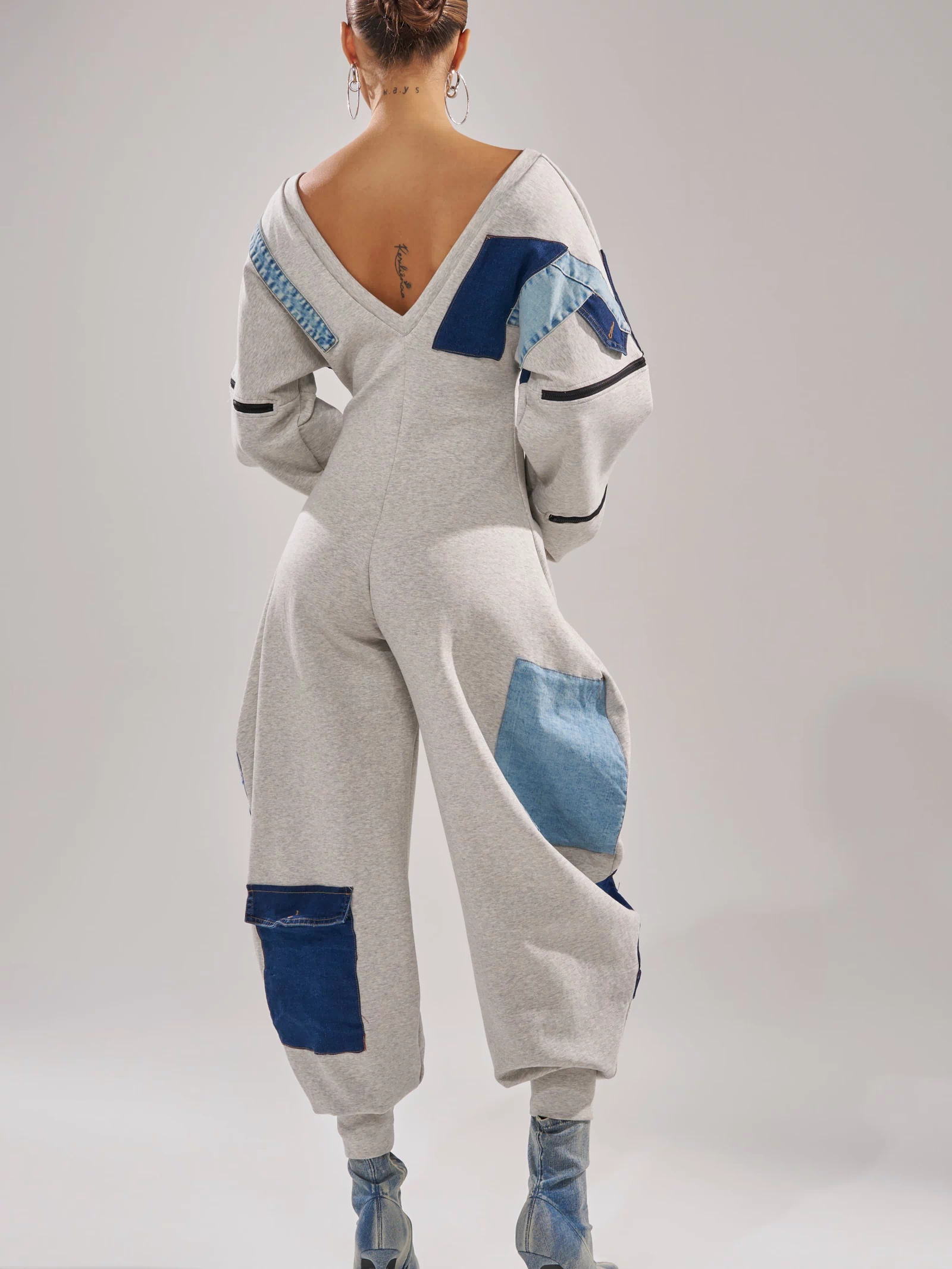 A person faces away in the COMFY COZY PATCHWORK JUMPSUIT IN HEATHER GREY with a deep V-back. Their hair is styled in a bun with large hoop earrings and blue heeled boots, creating a chic, modern streetwear one-piece ensemble.