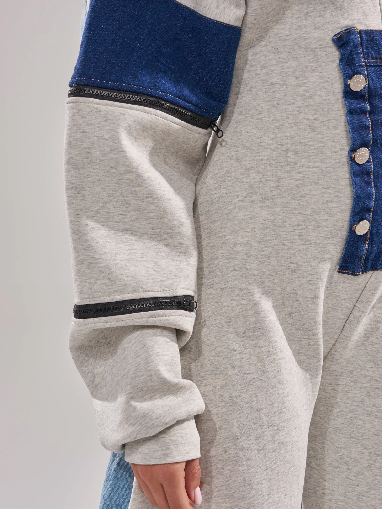 A person models the COMFY COZY PATCHWORK JUMPSUIT IN HEATHER GREY, featuring a blue denim chest panel, silver buttons, and black zippers on the sleeve. Only their lower face, torso, and part of the arm are visible.