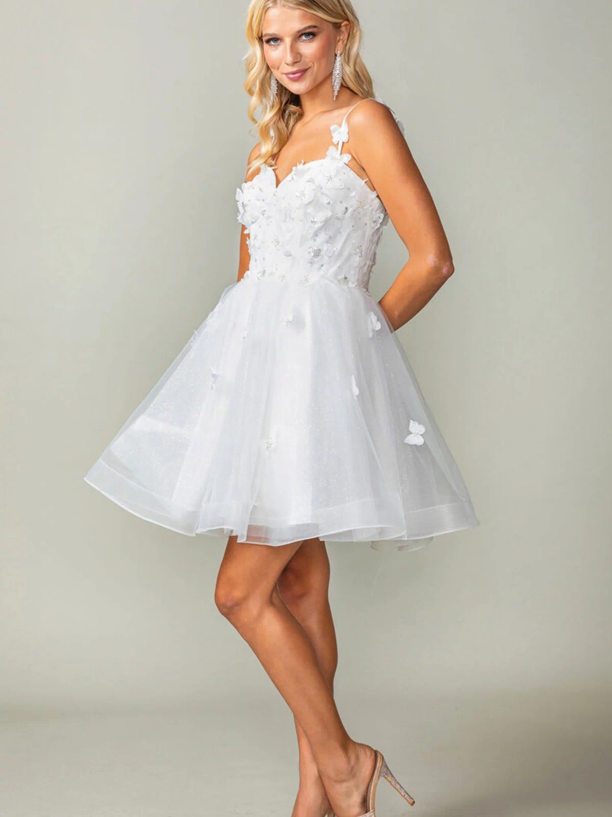 A woman in the Lace-Up Short Butterfly Homecoming Dress 3369, a white semi-formal dress with delicate 3-D butterflies, and high heels stands against a plain light background.